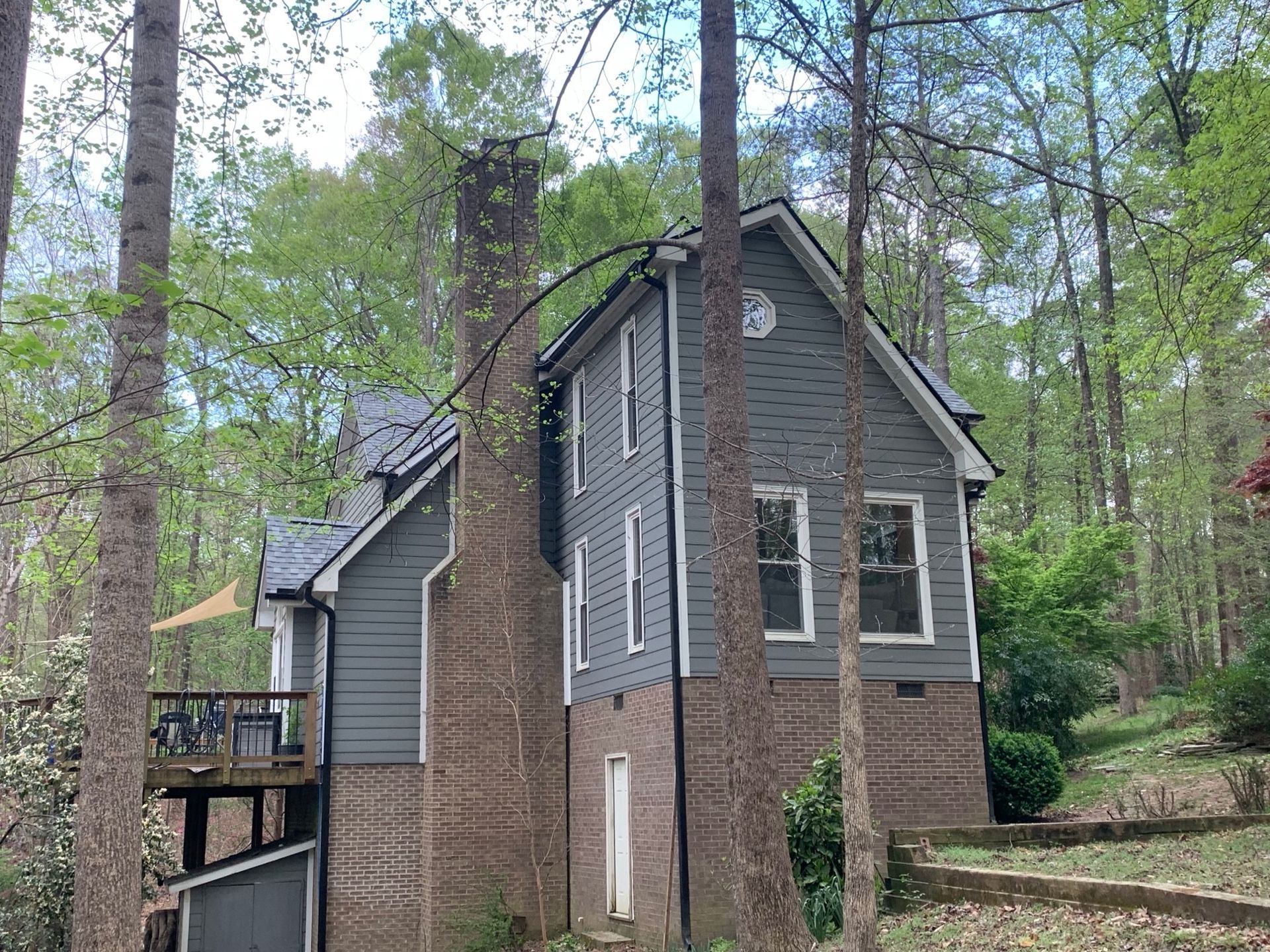 Gray house with brick chimney, nestled among trees, on a hillside.