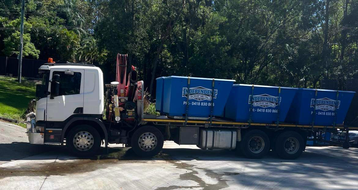 White truck with a flatbed carrying three blue bins on a paved road, trees in the background— Independent Scrap Metal Pty Ltd in Yamba, NSW