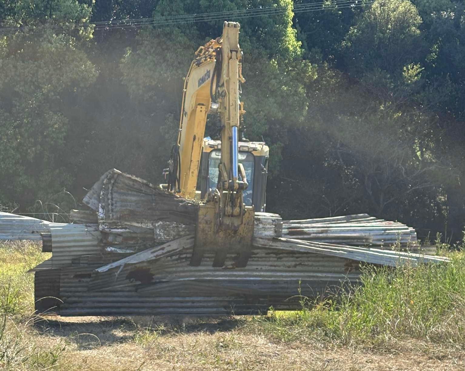 Yellow excavator lifting and moving sheets of metal. Outdoors, with trees in background— Independent Scrap Metal Pty Ltd in South Lismore, NSW