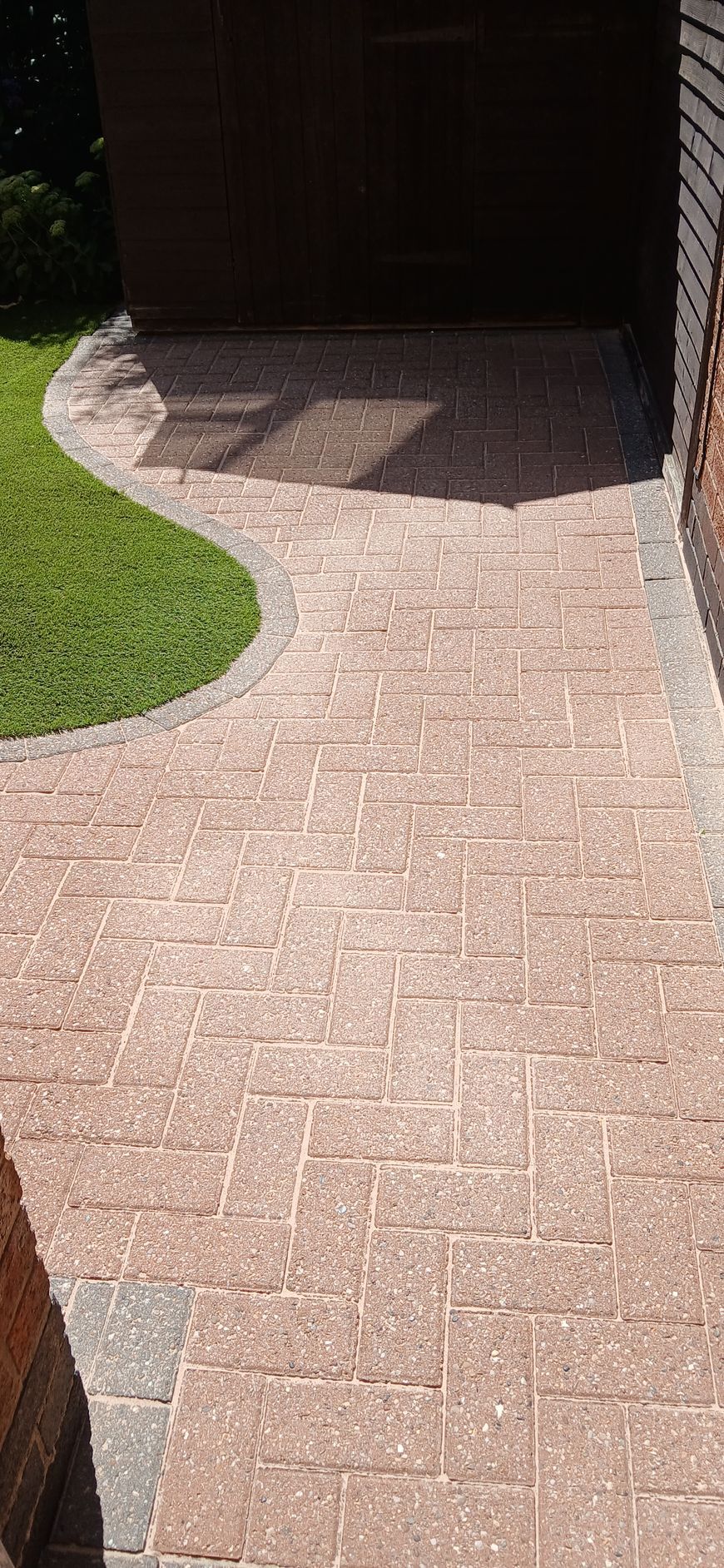 A block paved patio leading to a shed next to a lawn area after being pressure washed.