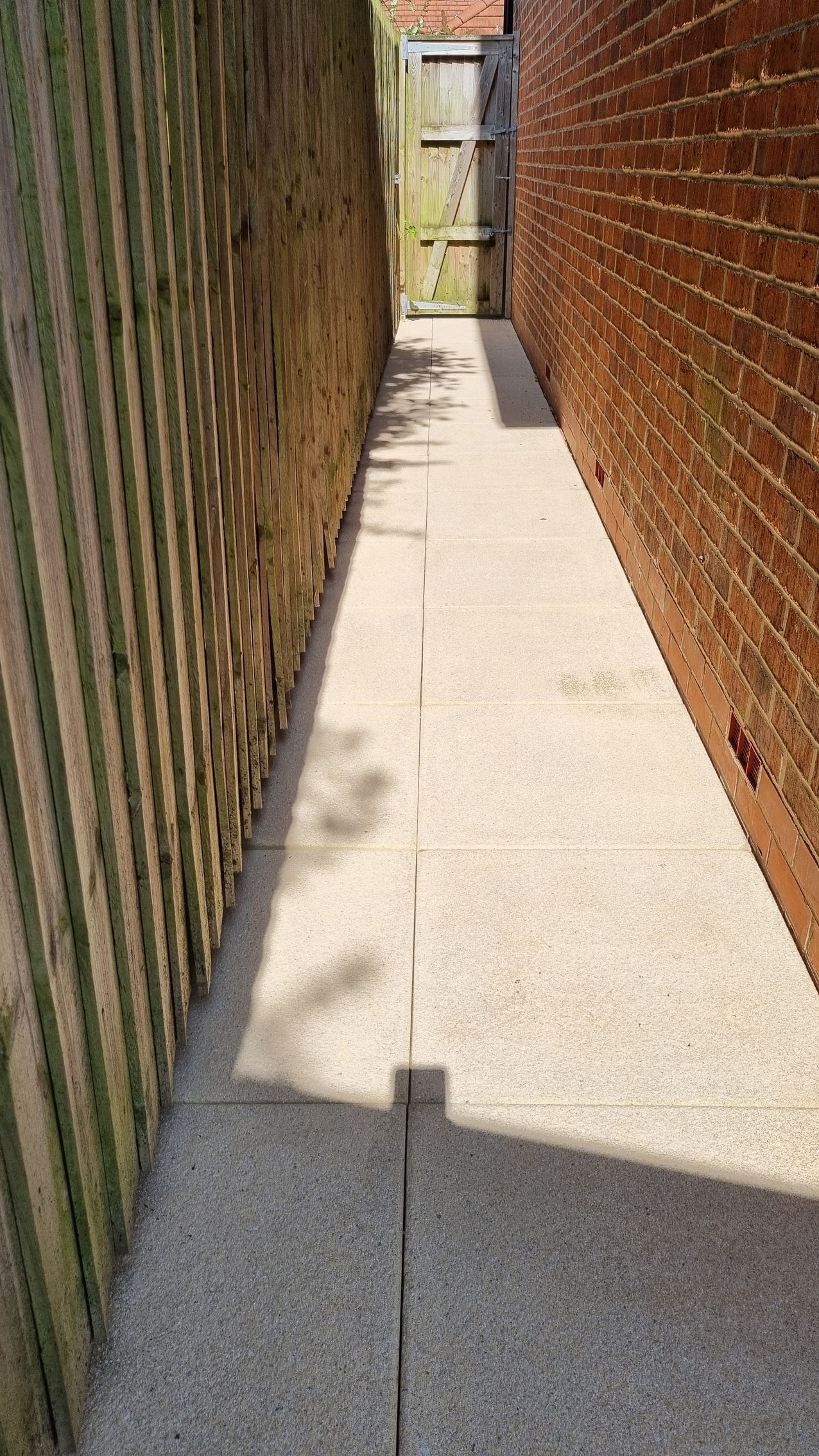 A footpath /patio between a house and wooden fence that has recently been pressure washed.