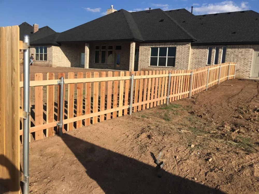 Wooden picket fence in a yard, extending toward a brick house with dark roof.