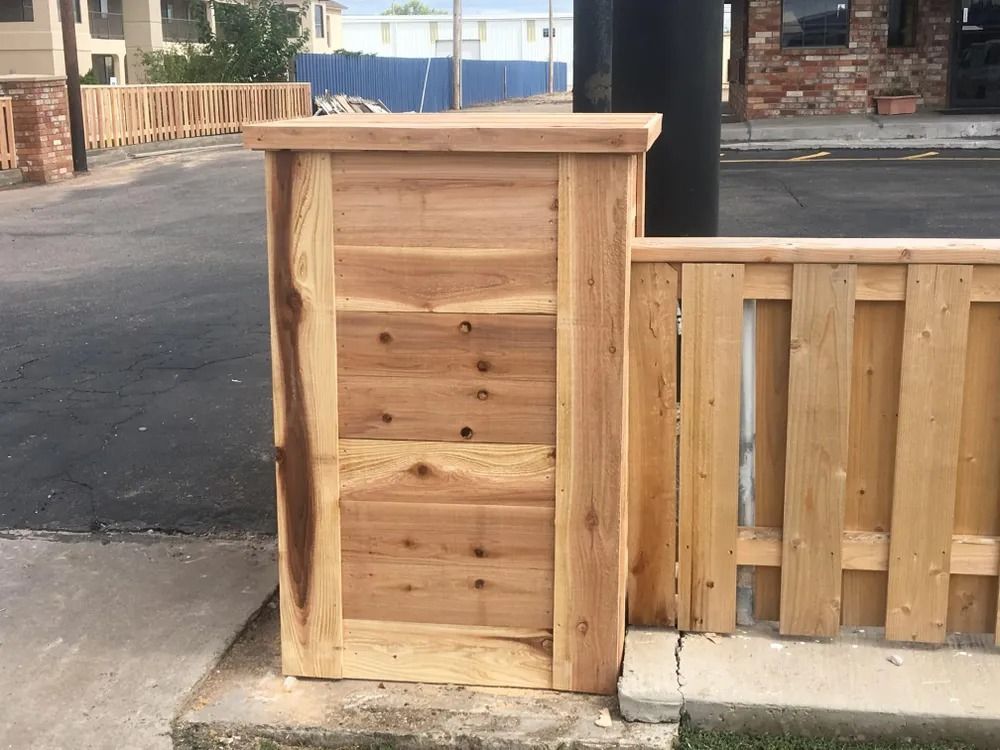 Wooden pillar next to a wooden fence on concrete, with road and building visible.
