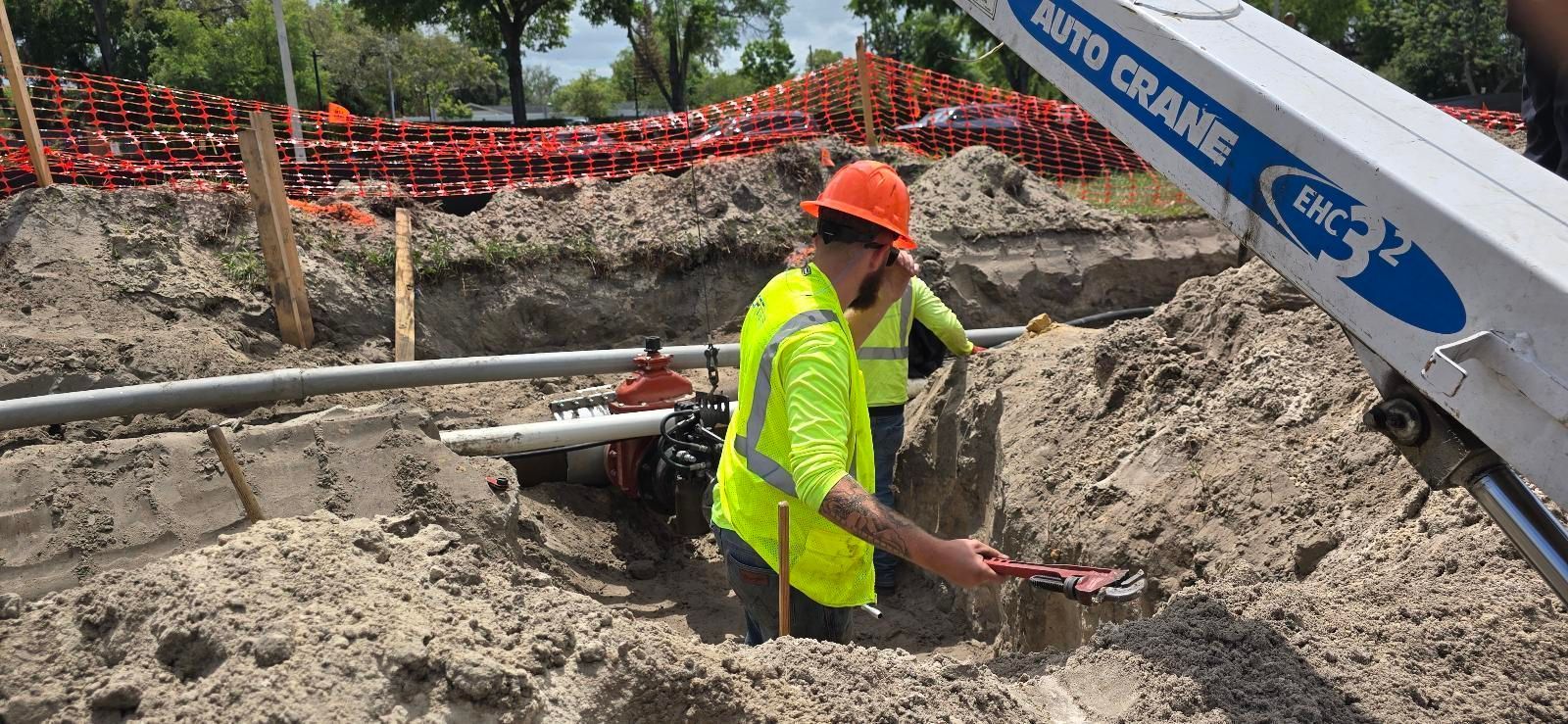 Construction workers at a site with an excavator and pipes. One man stands, two others work on the pipes.