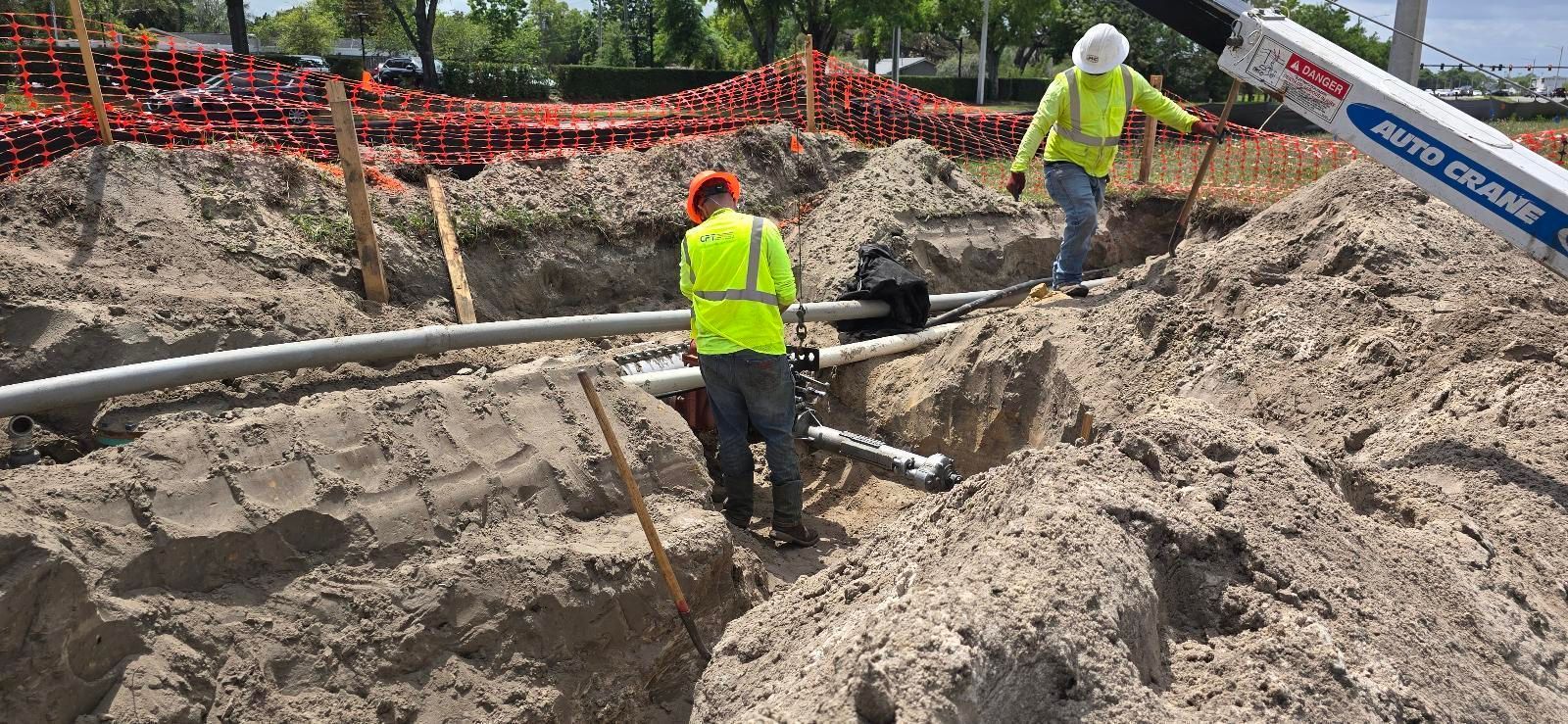 Two workers by an exposed pipe in an excavation, assisted by an excavator. Workers wear safety vests.