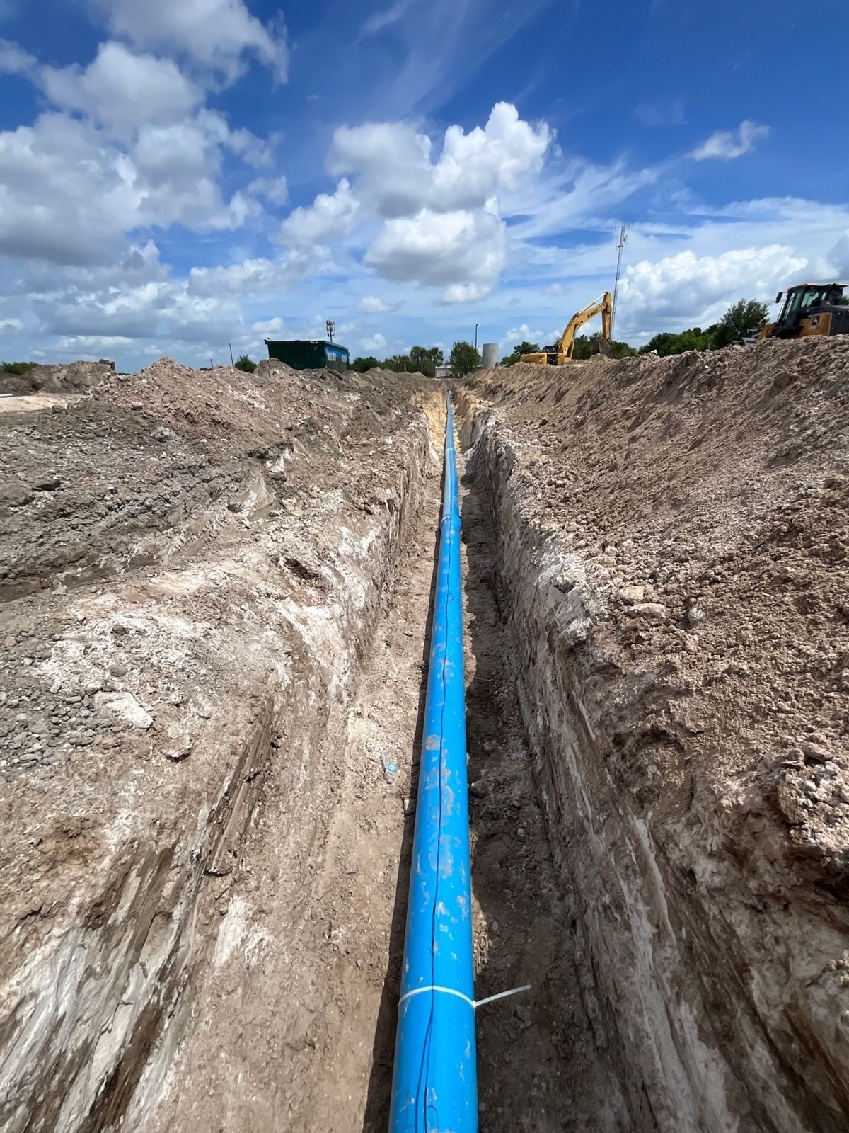 Construction site: Person using jackhammer on sidewalk, concrete debris, white fence, and vehicles in background.