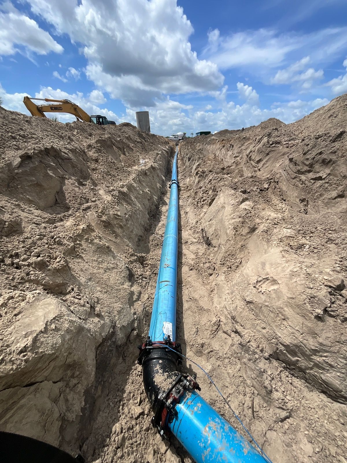 Workers in a trench connecting a large blue pipe with an excavator nearby, sunny outdoor setting.