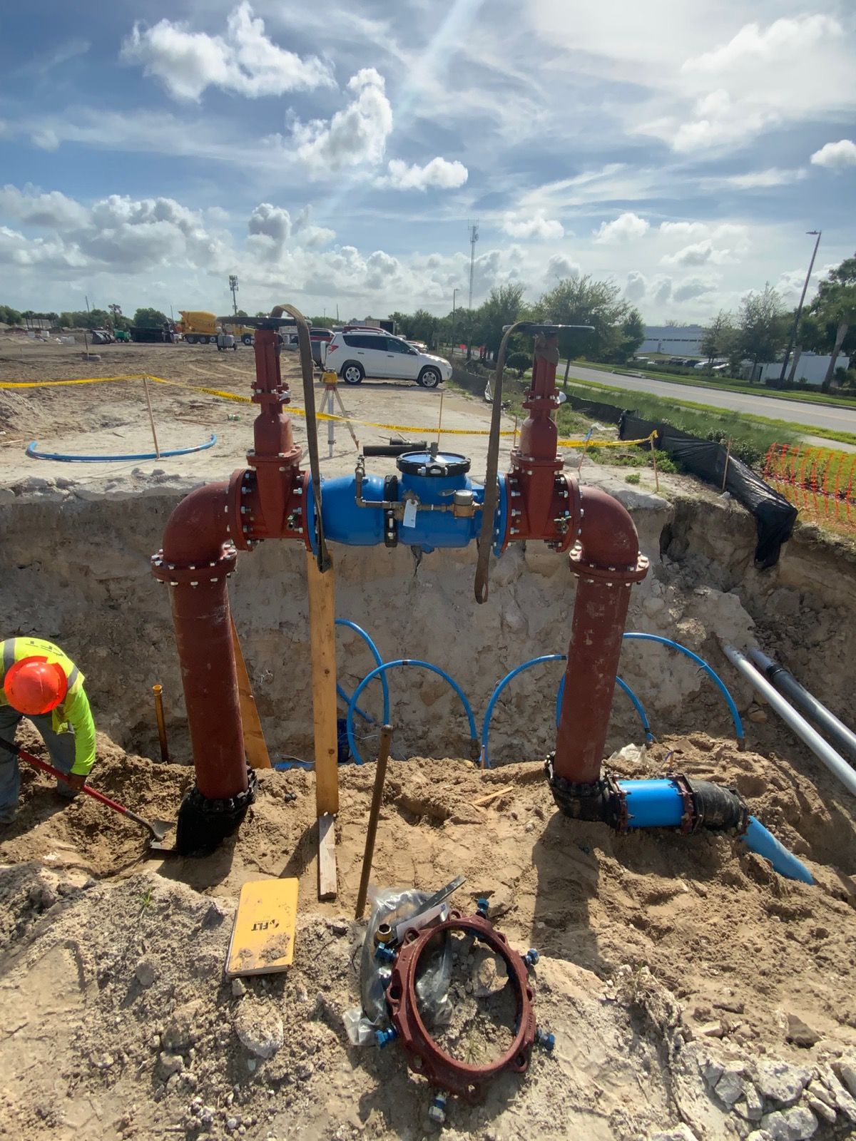 Construction worker in a safety vest near excavator and black pipe in a dirt pit. Sunny day, industrial setting.