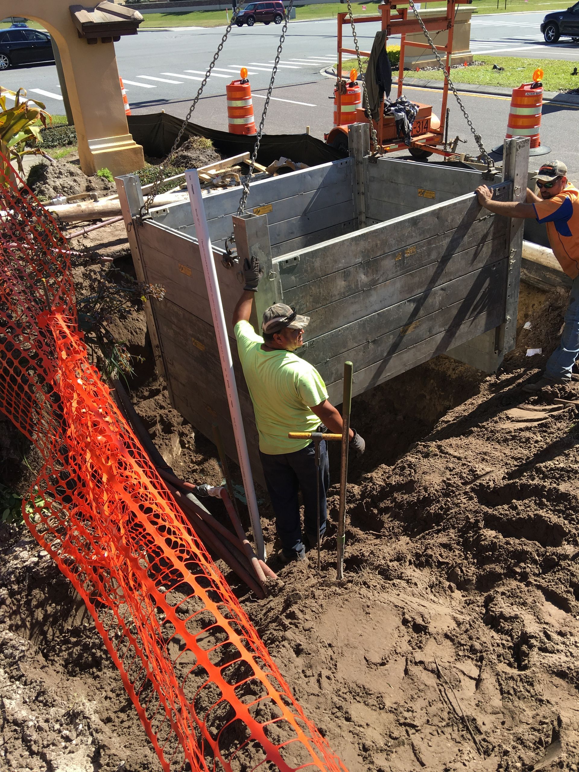 Workers in a trench, lowering a metal shoring box. Orange safety fence and cones are in the background.