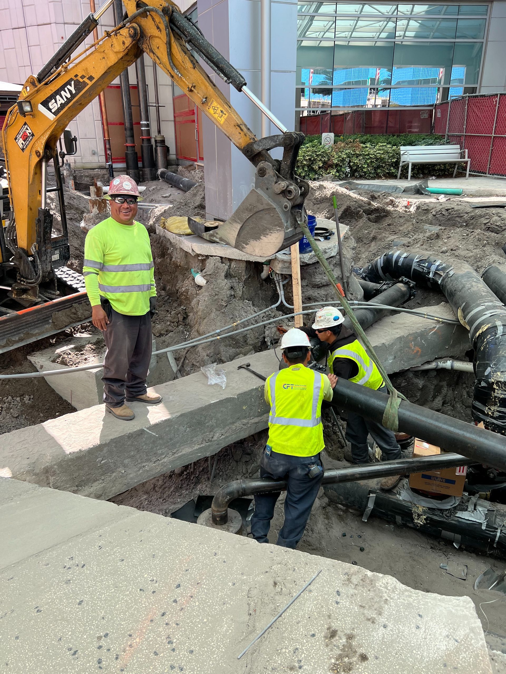 Construction workers at a site with an excavator and pipes. One man stands, two others work on the pipes.