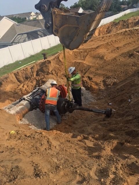 Two workers by an exposed pipe in an excavation, assisted by an excavator. Workers wear safety vests.