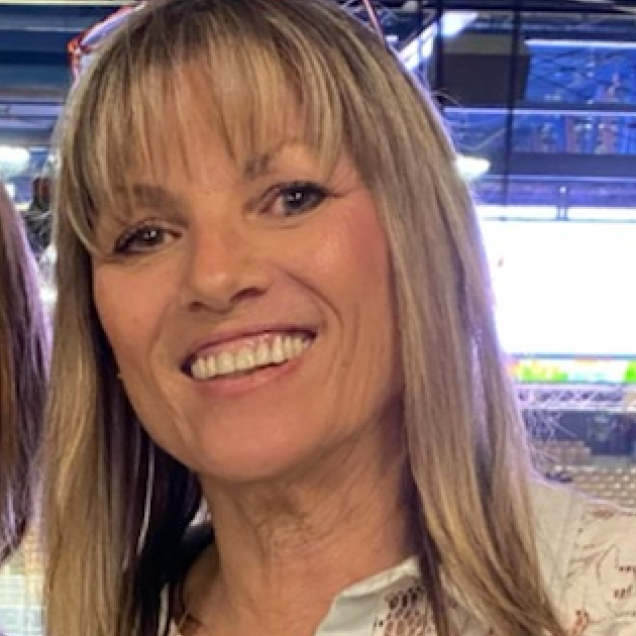 Woman with blonde hair smiling, possibly indoors, with a blurry background.