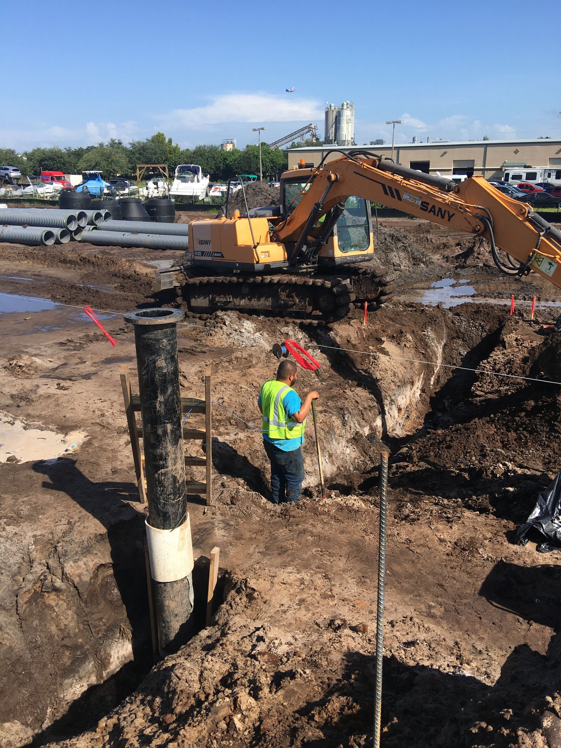 Construction worker in a safety vest near excavator and black pipe in a dirt pit. Sunny day, industrial setting.