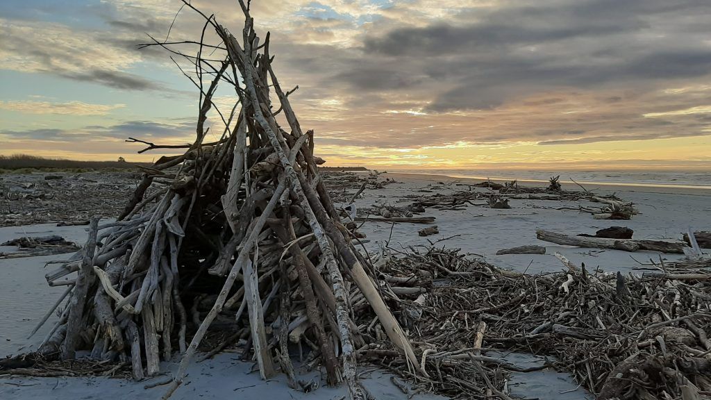 A hut made out of driftwood on a beach at sunset.