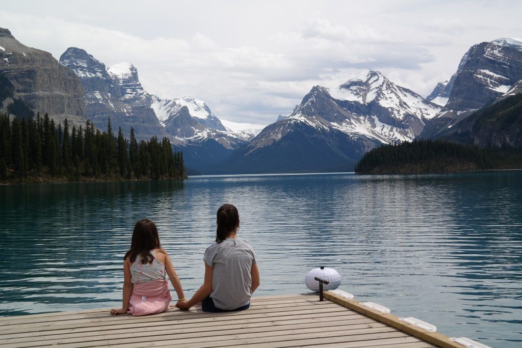 Two girls are sitting on a dock looking at a lake with mountains in the background.