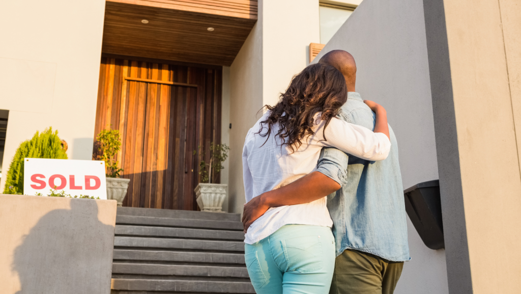 A man and woman are standing in front of a house with a sold sign.