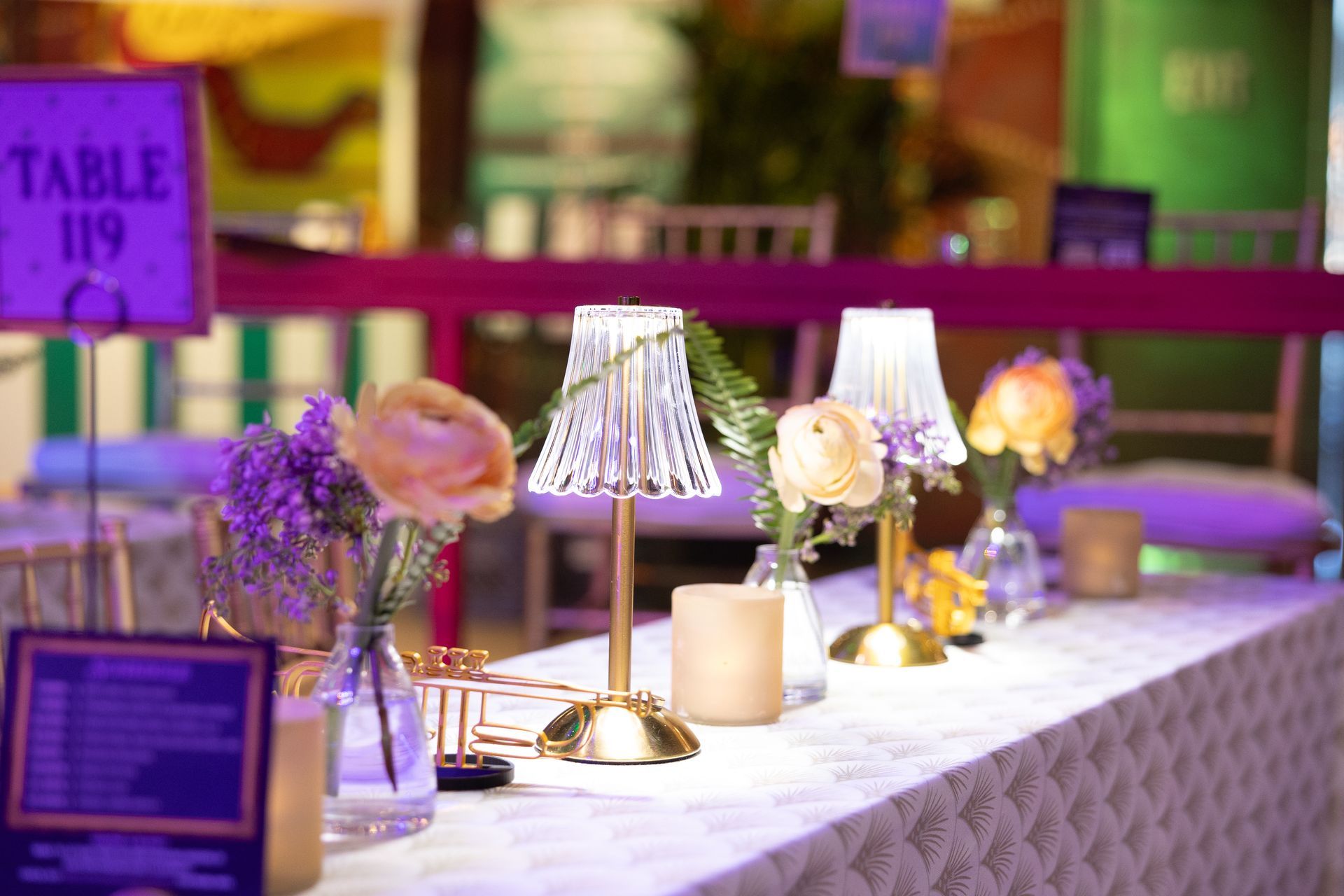 Table setting with flowers, lamps, and table number 119 at a party. Gold, pink, and purple colors.