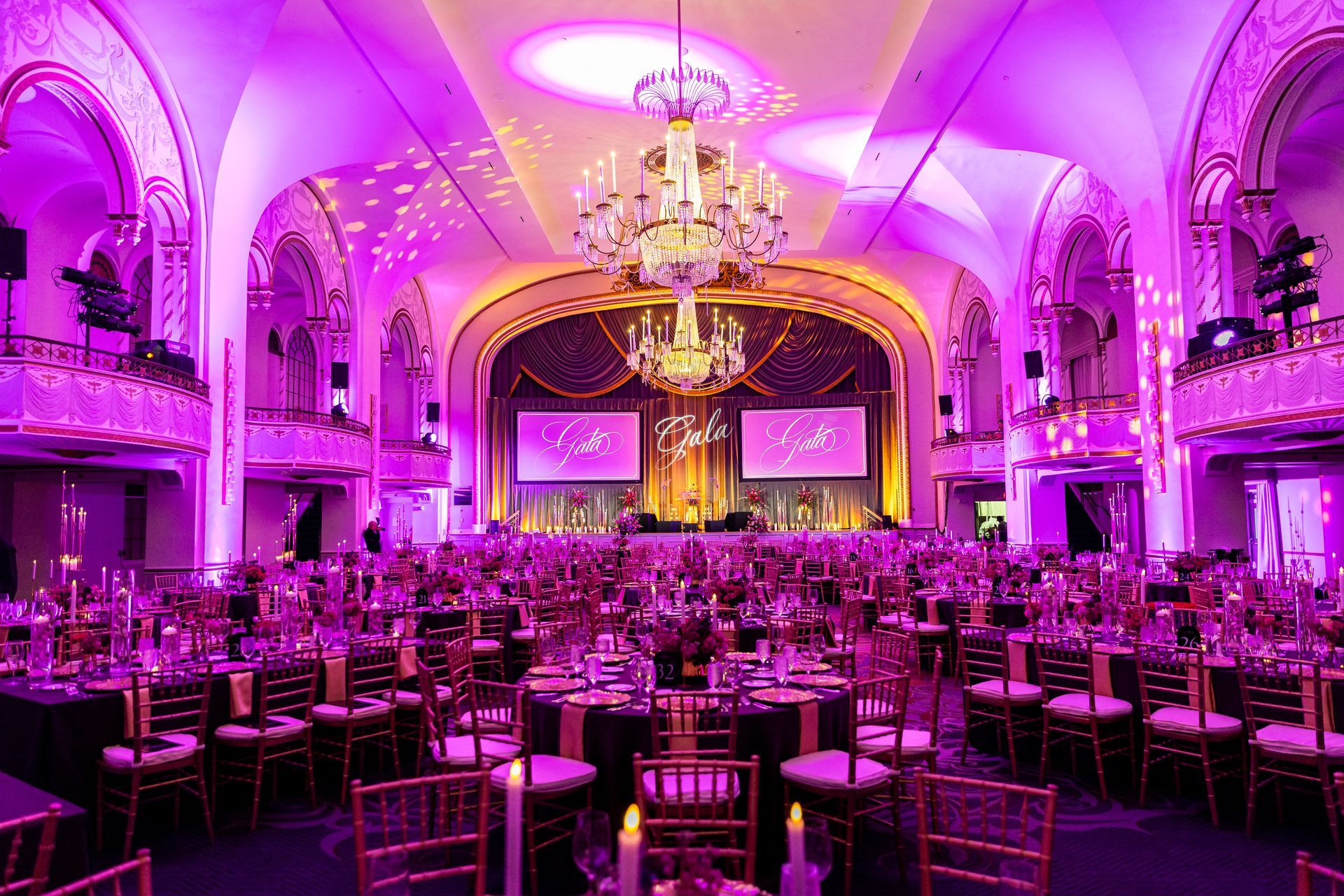 Ornate ballroom set for an event, lit in pink. Tables are set with chairs, and stage is at the back.