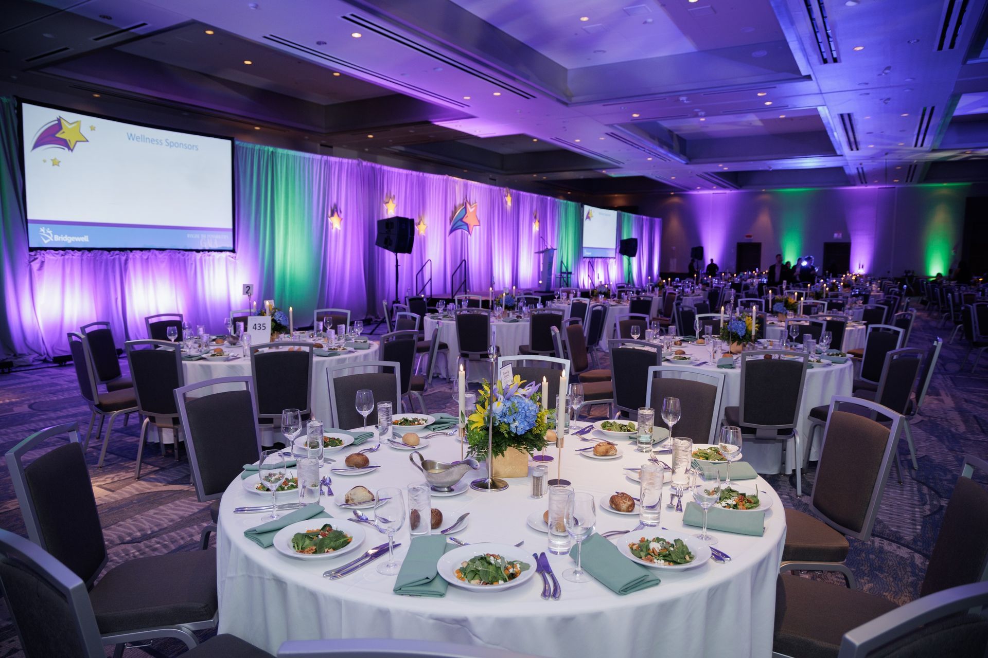 Banquet hall with round tables set for a formal event, lit with purple and green hues, and two screens.