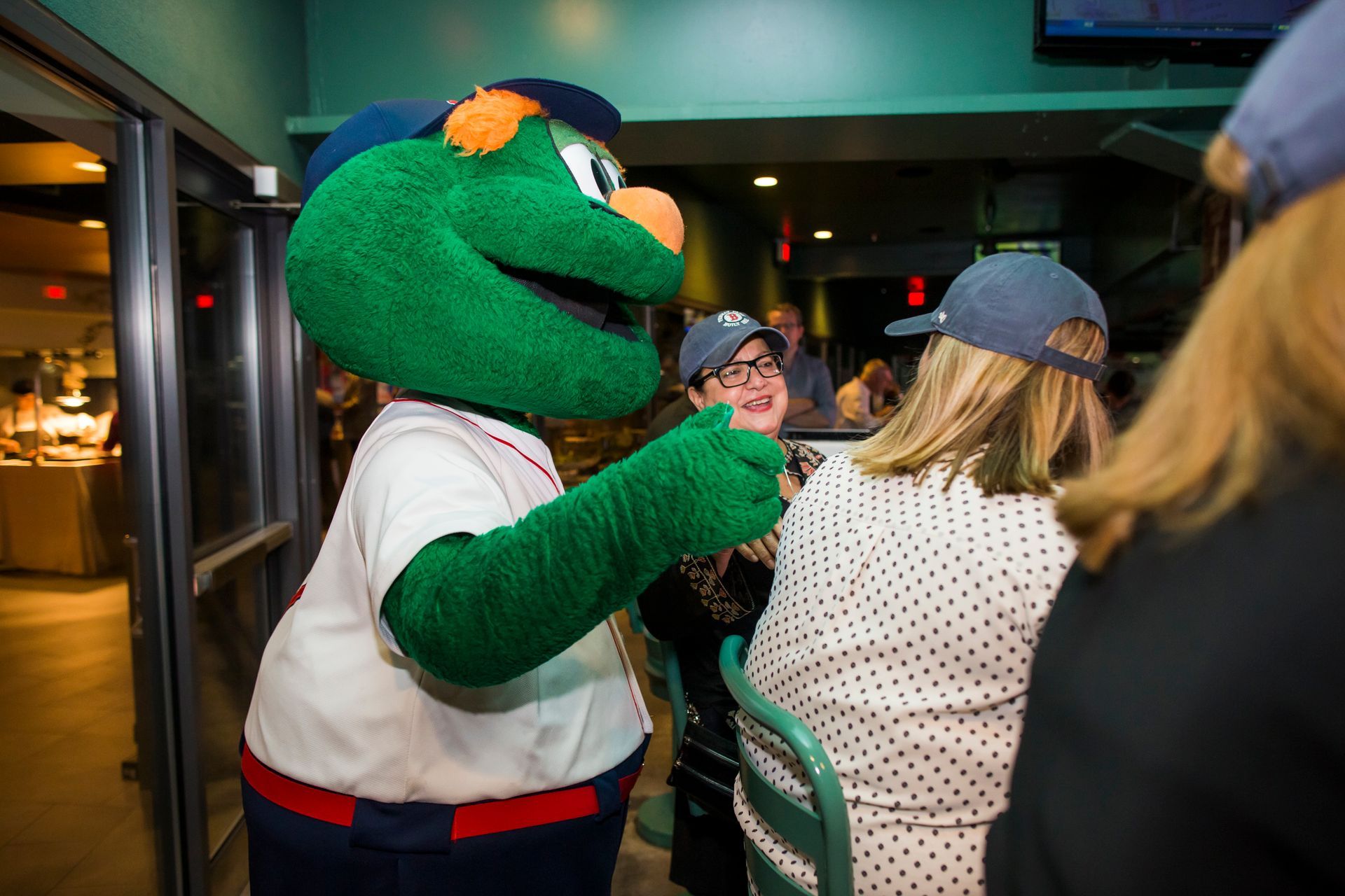Mascot in baseball uniform fist-bumping a woman in a restaurant, other people seated nearby.