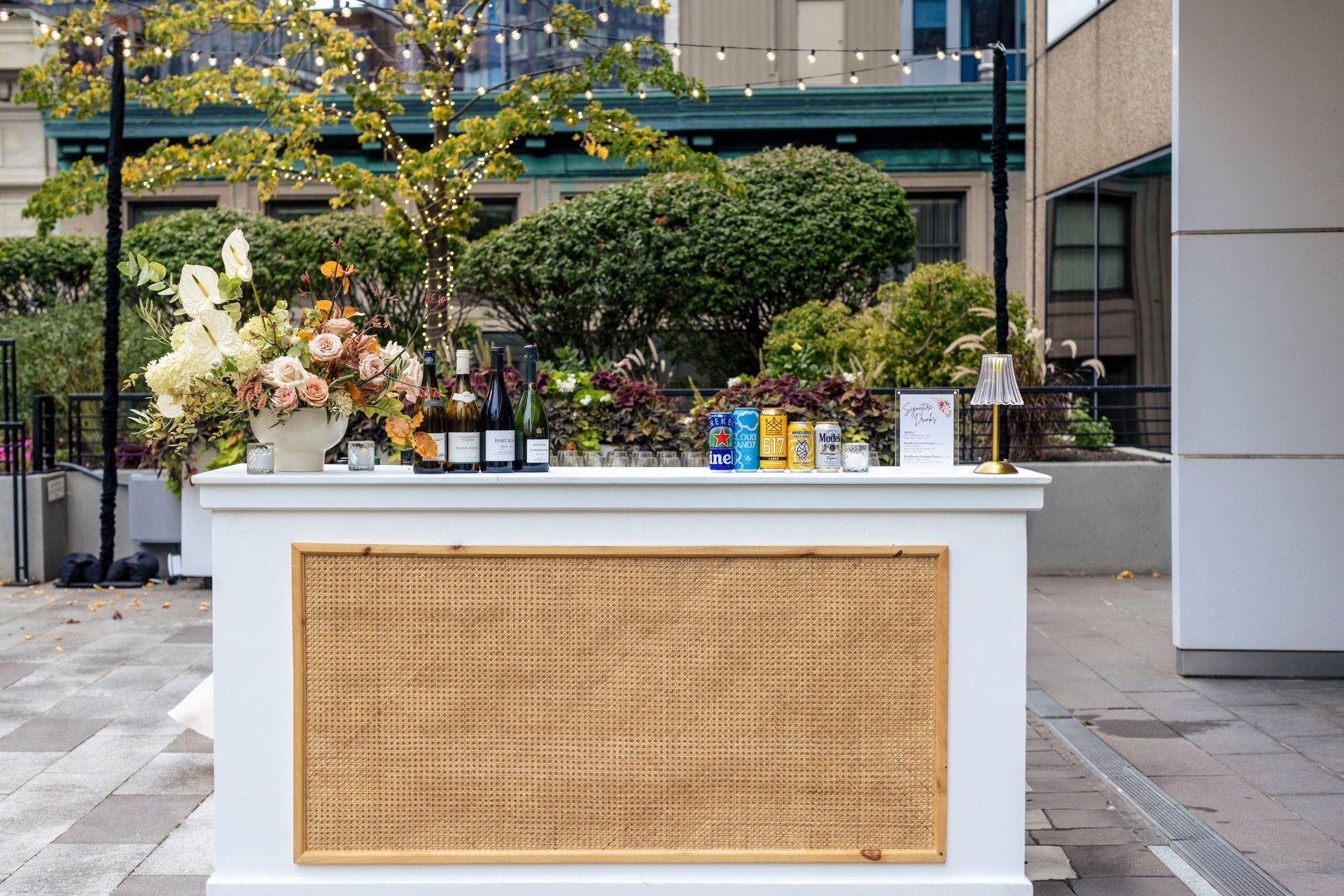 Outdoor bar with drinks and flowers; white bar, woven front, green foliage background.