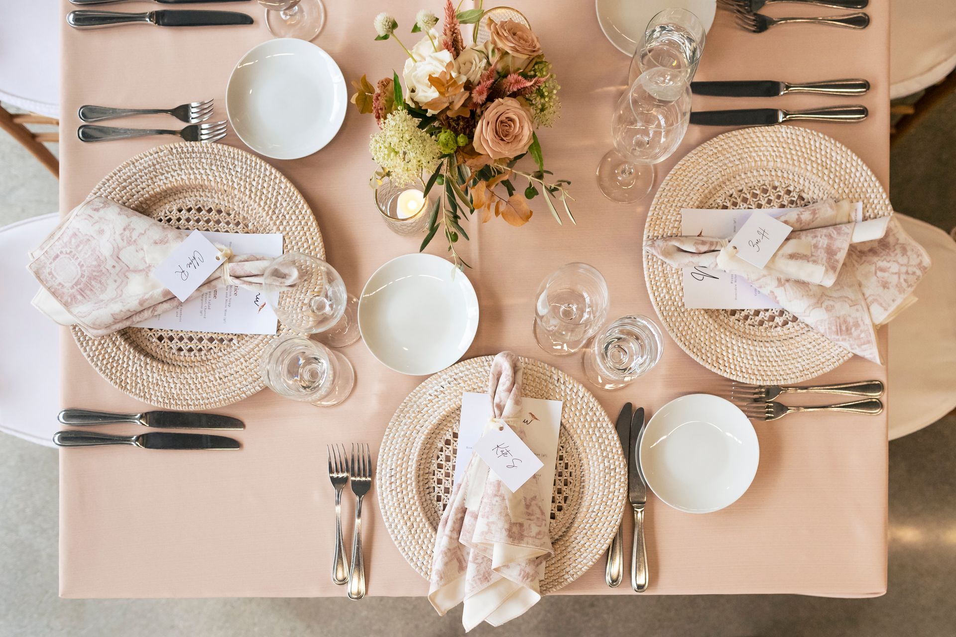 Elegant table setting with blush tablecloth, woven placemats, white dishes, floral centerpiece, and place cards.