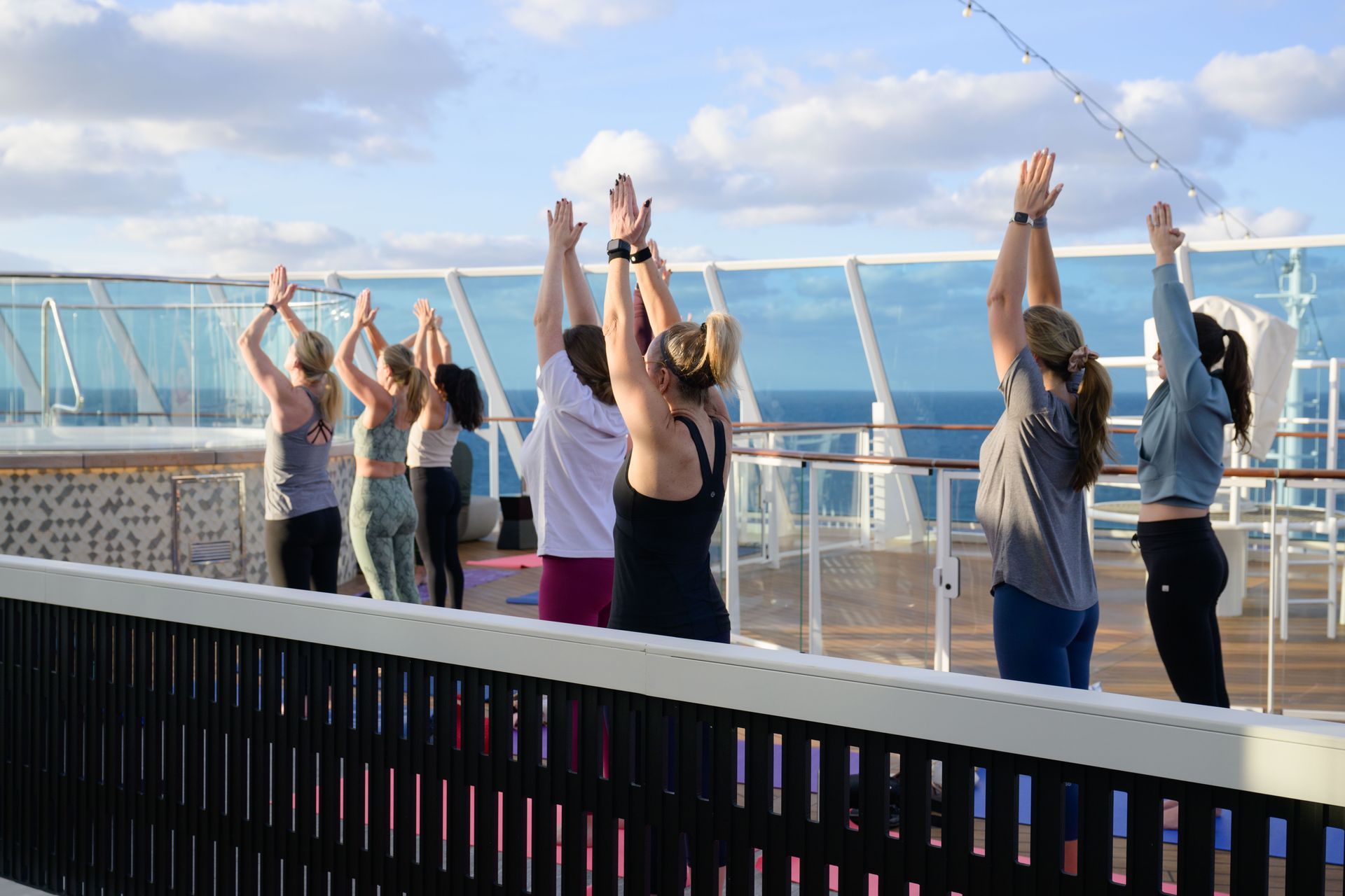 People doing yoga on a cruise ship deck, arms raised, with ocean view and blue sky.