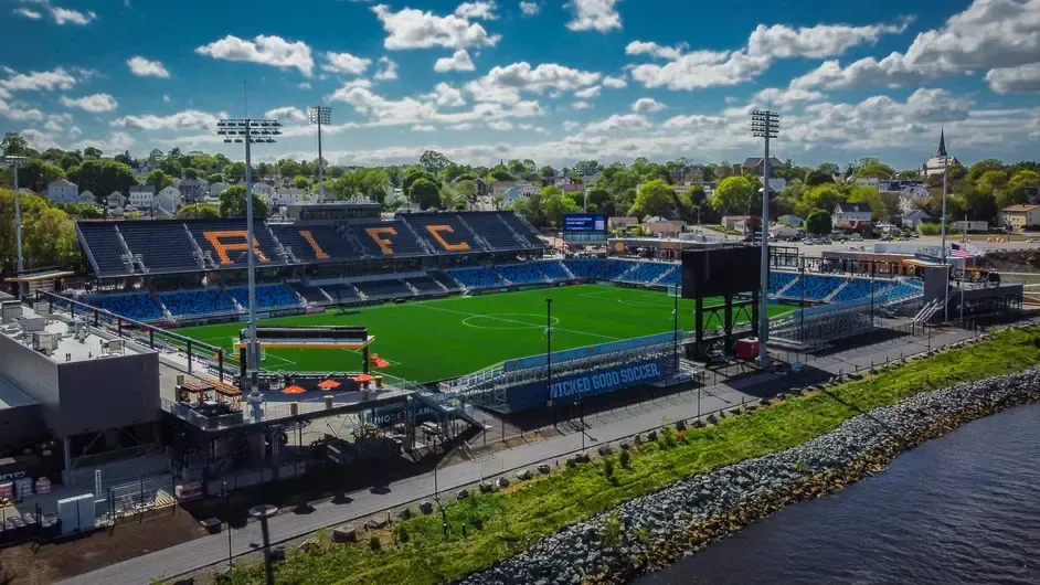 An aerial view shows a soccer stadium. Blue seats, green field, and the letters
