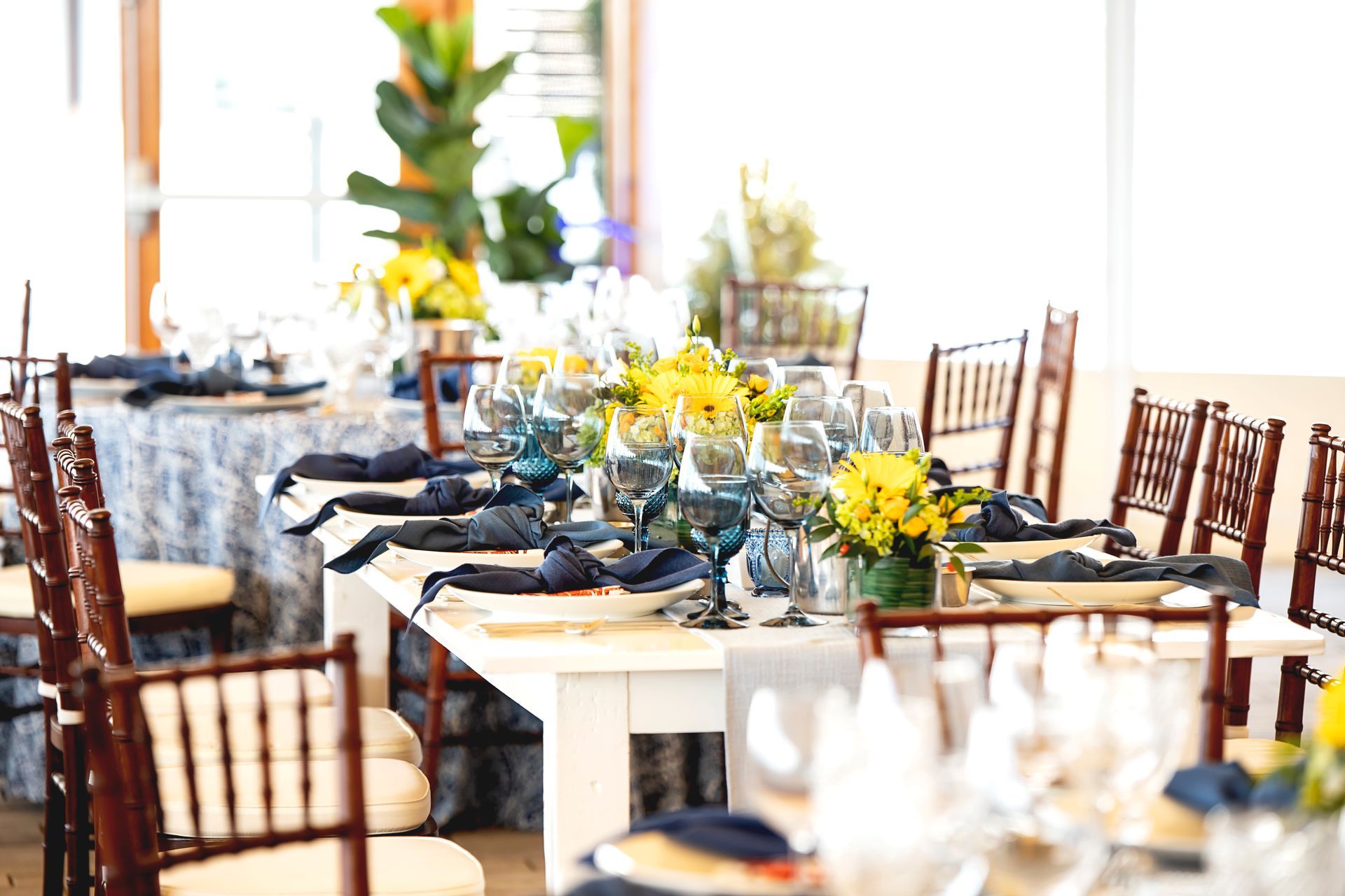 Long dining table set for a formal event with blue glassware, yellow flowers, and dark blue napkins.