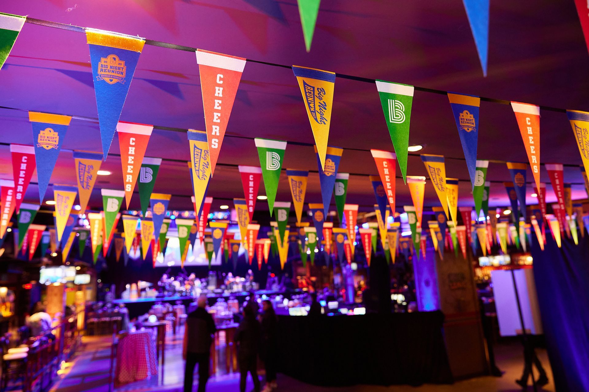 Colorful pennant flags hang over a venue with people, tables, and stage lighting.