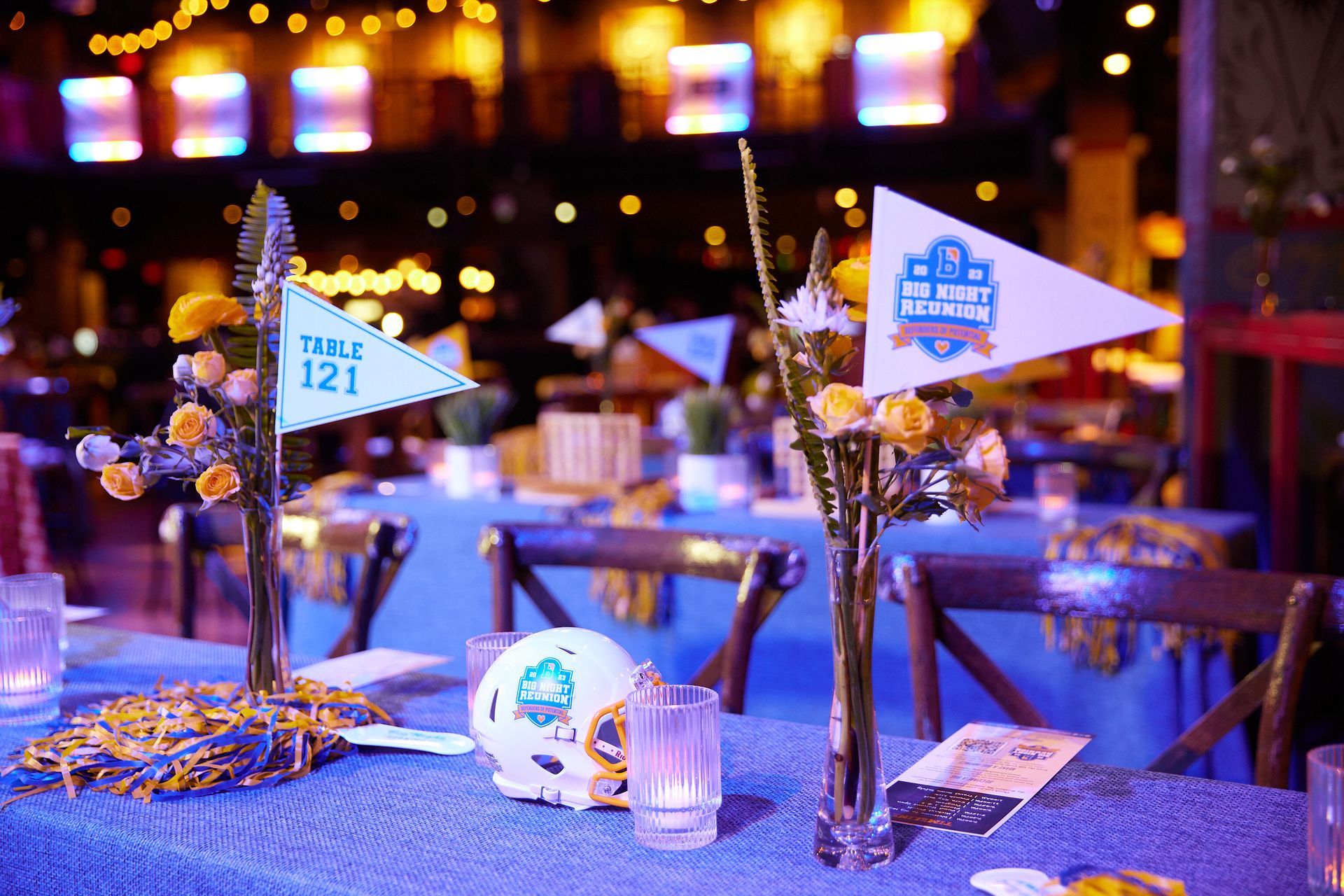 Table setting at a sports-themed event with blue tablecloth, flags, flowers, and centerpieces.