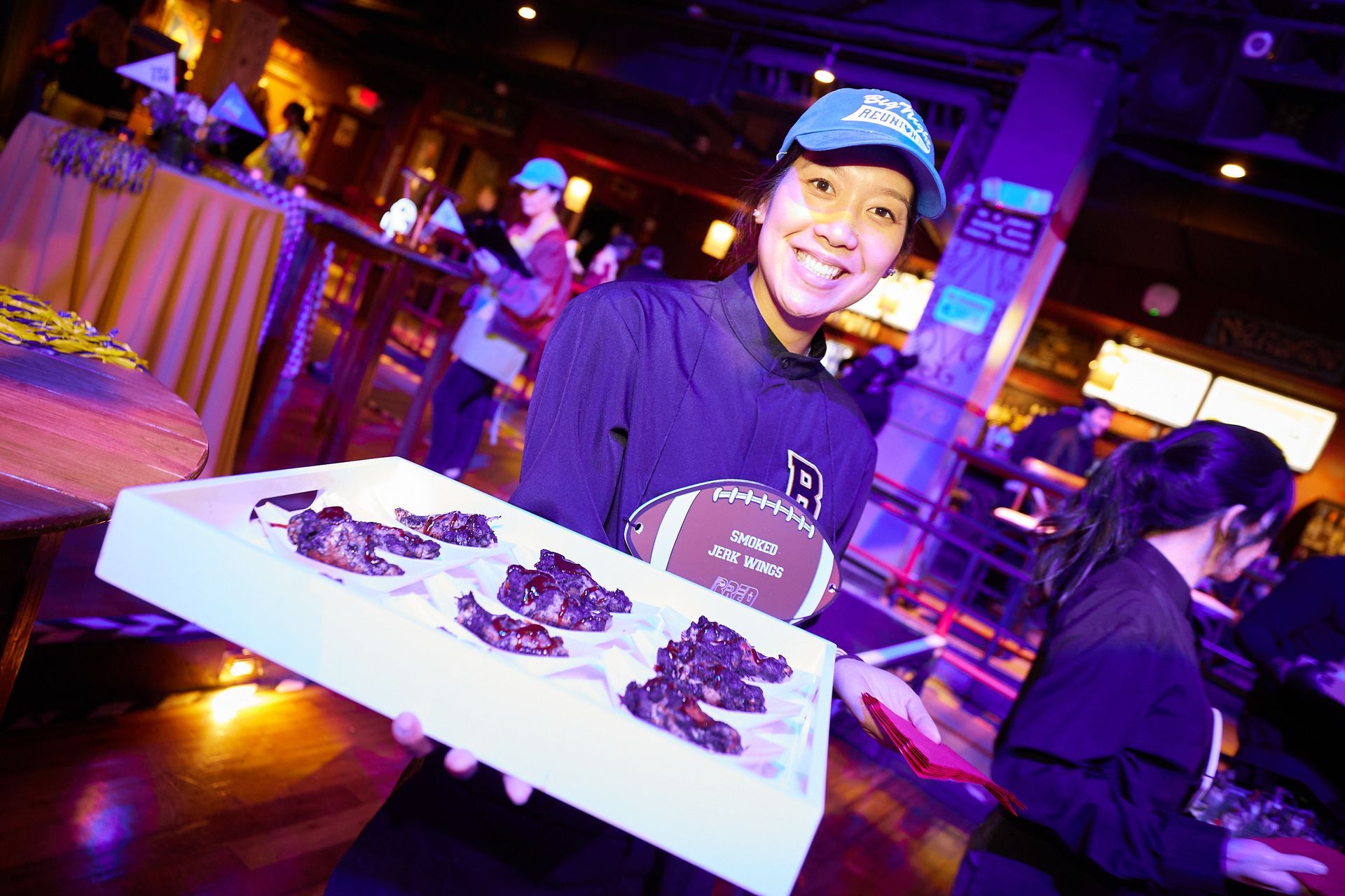 Woman in cap smiling, holding a tray of food at a party. Wooden interior, other people in background.