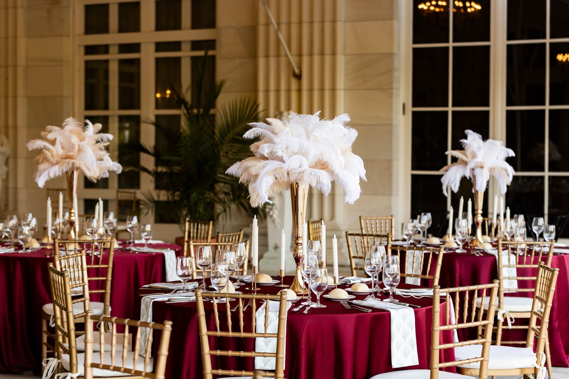 Elegant dining tables set with maroon tablecloths, gold chairs, and white feather centerpieces outdoors.