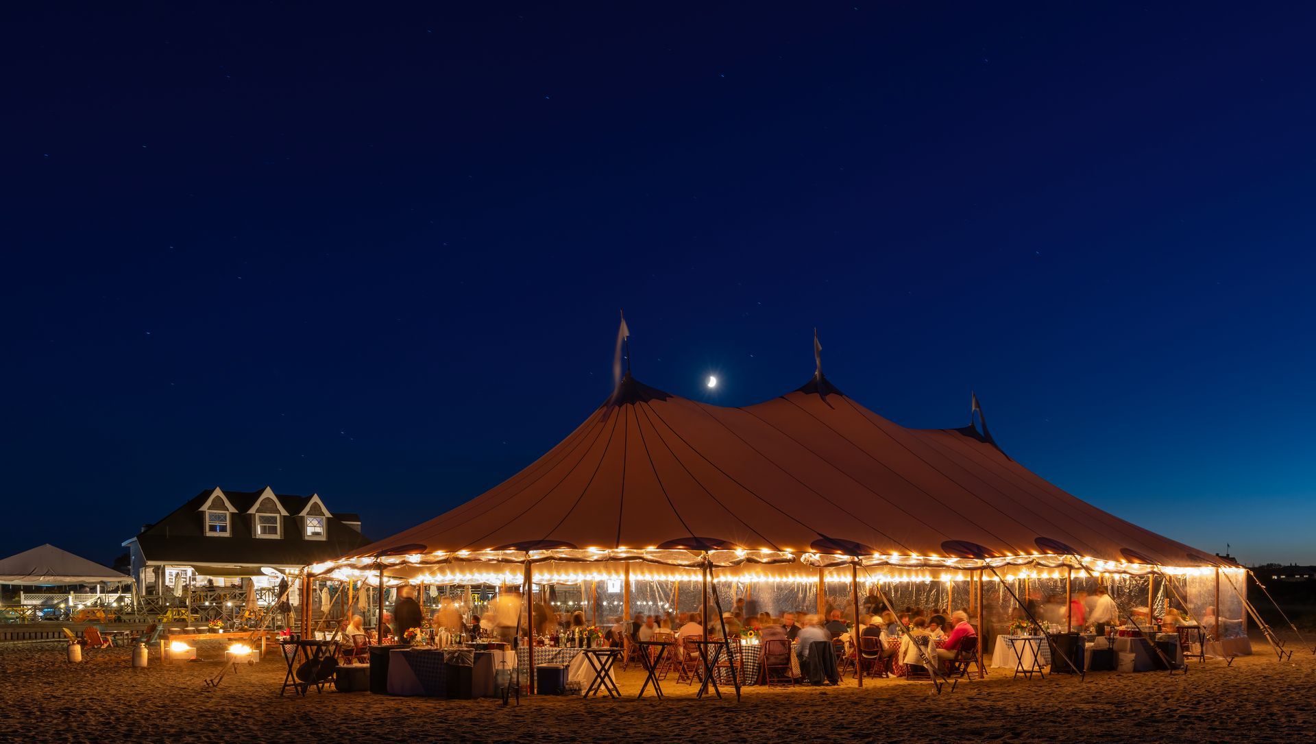 A large, lit tent at night, with people dining, next to a building with illuminated windows.