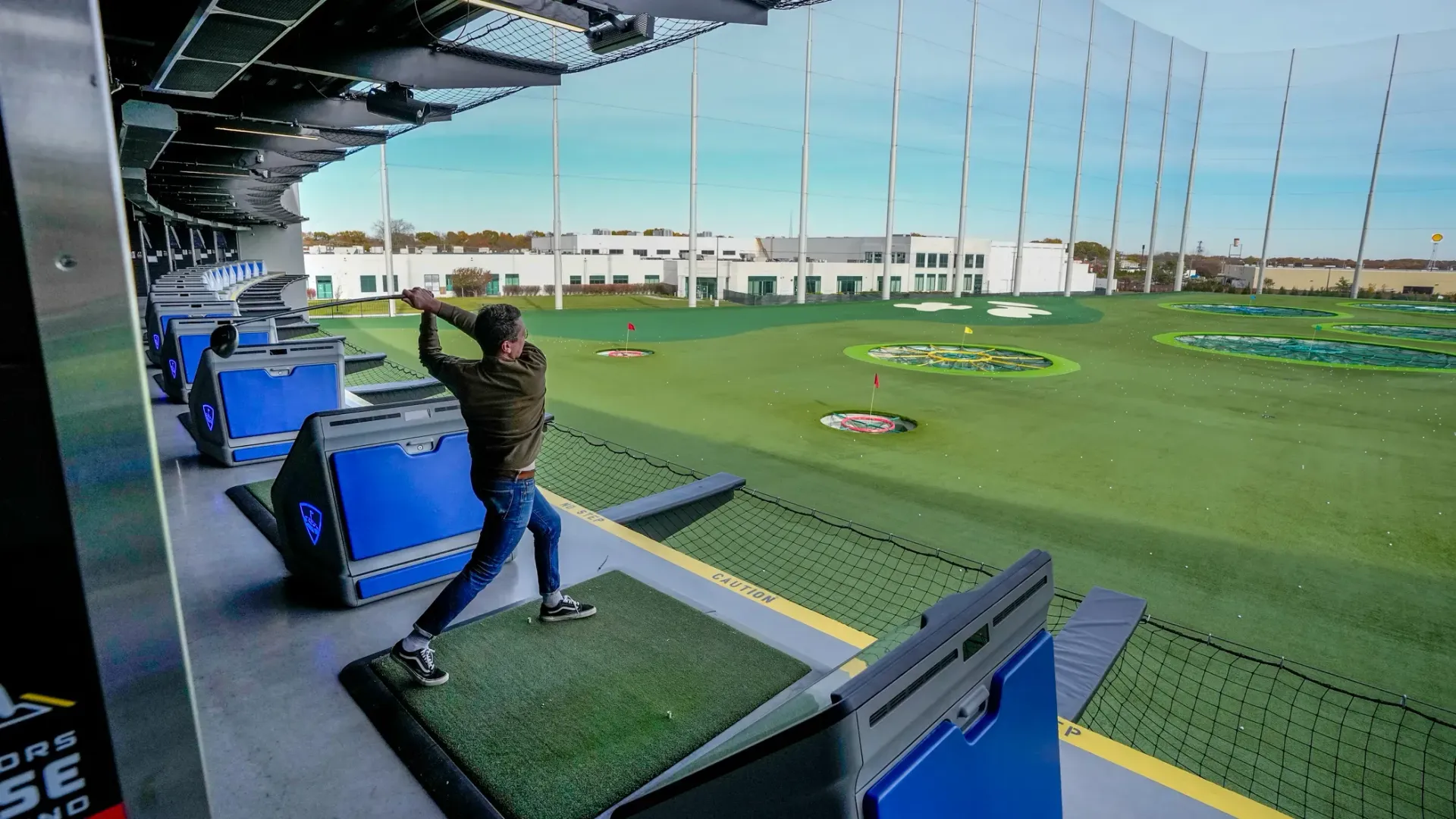 Person swinging a golf club at a Topgolf. Bright green field, blue bays, safety netting.