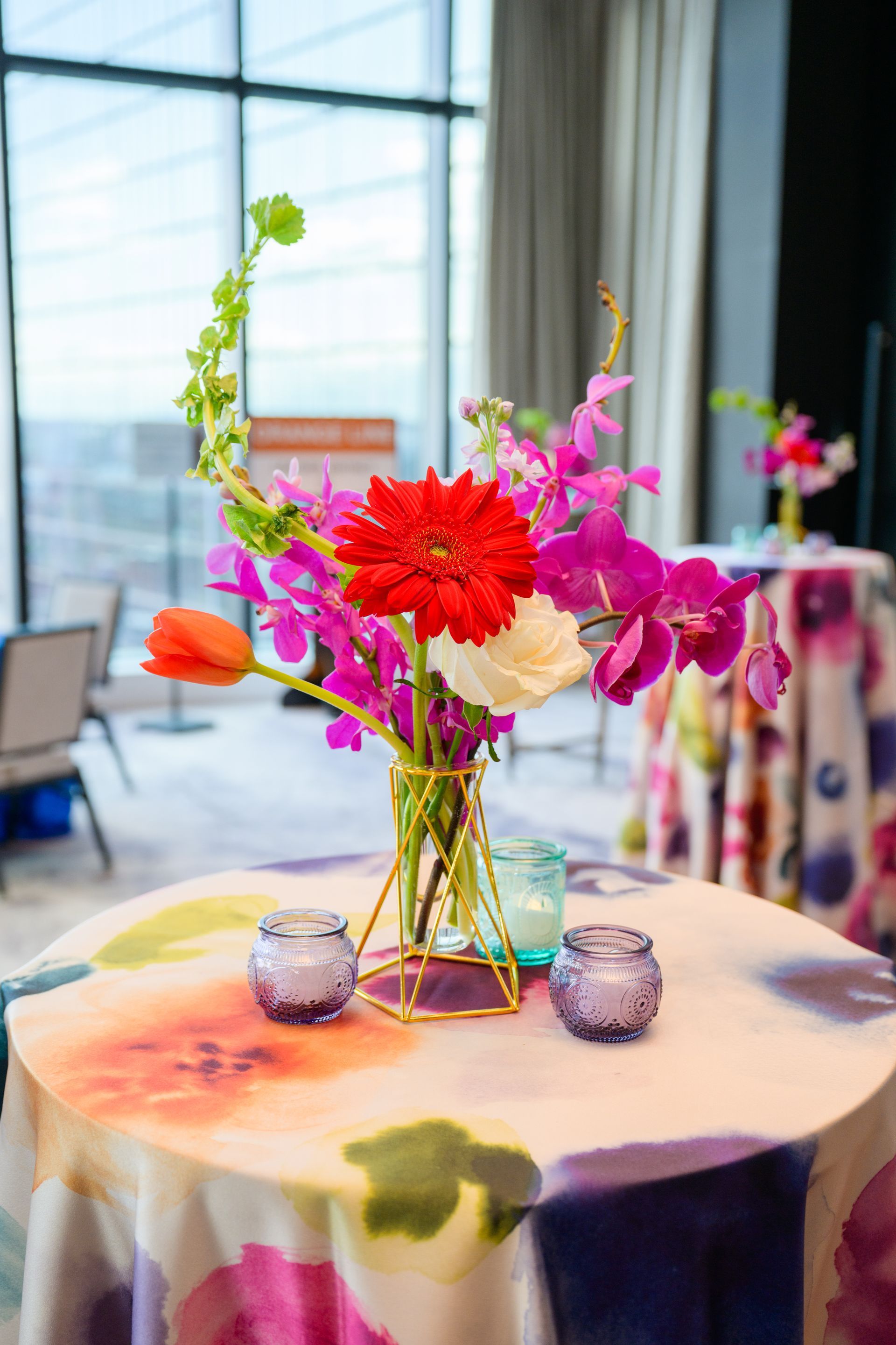 Floral centerpiece with red gerbera, pink orchids, and candles on a round table with floral tablecloth.