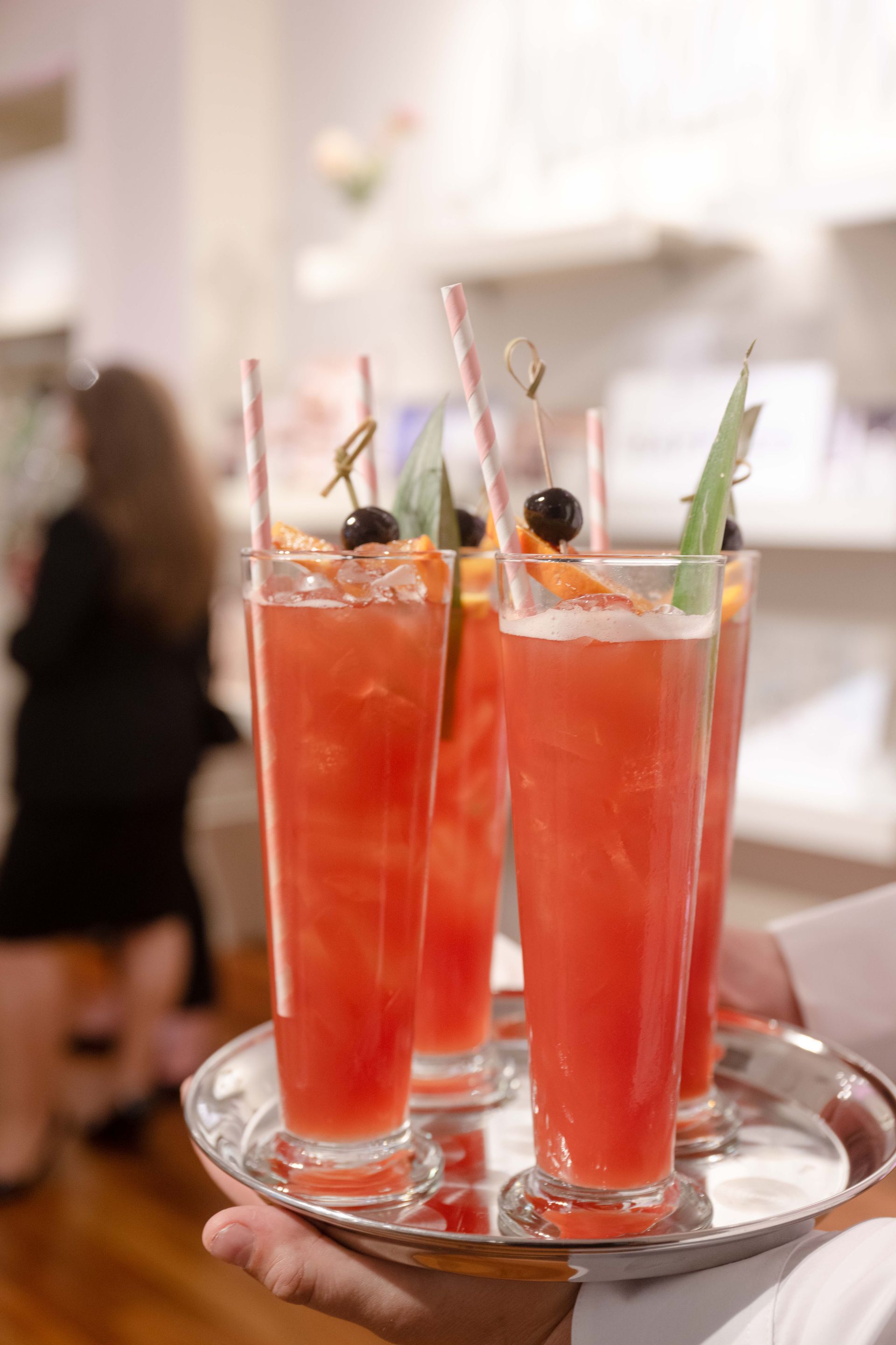 A server holding a silver tray with four tall, red cocktails garnished with fruit and straws.