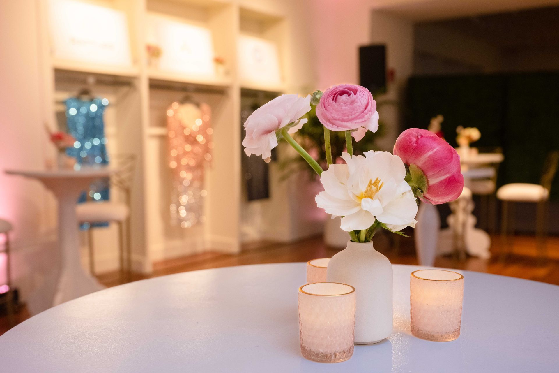 Flowers and candles on a round table in front of a clothing display; pink and white decor.