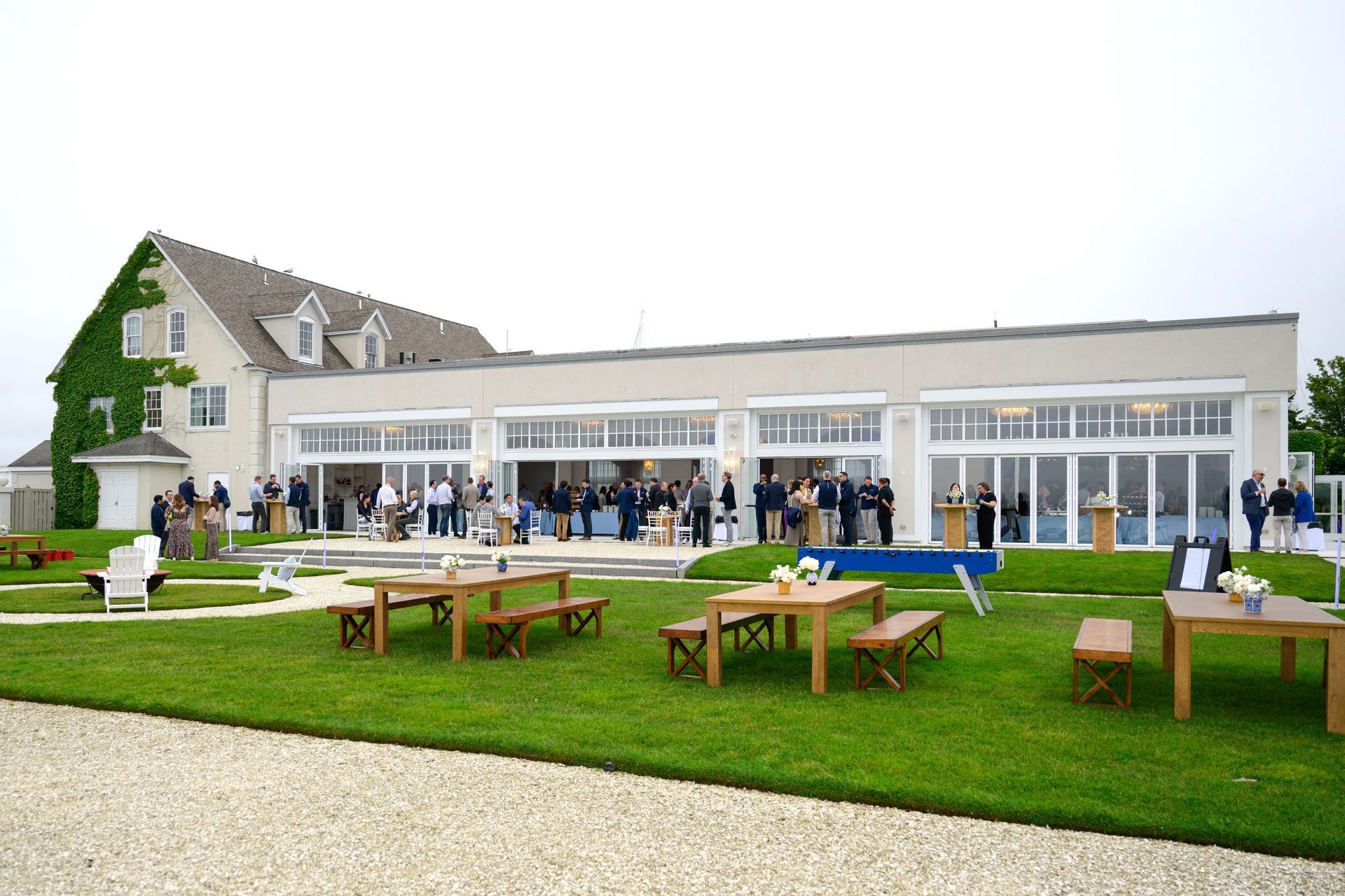 People gather for an outdoor event at a modern building with large glass doors and a grassy lawn featuring wooden tables.