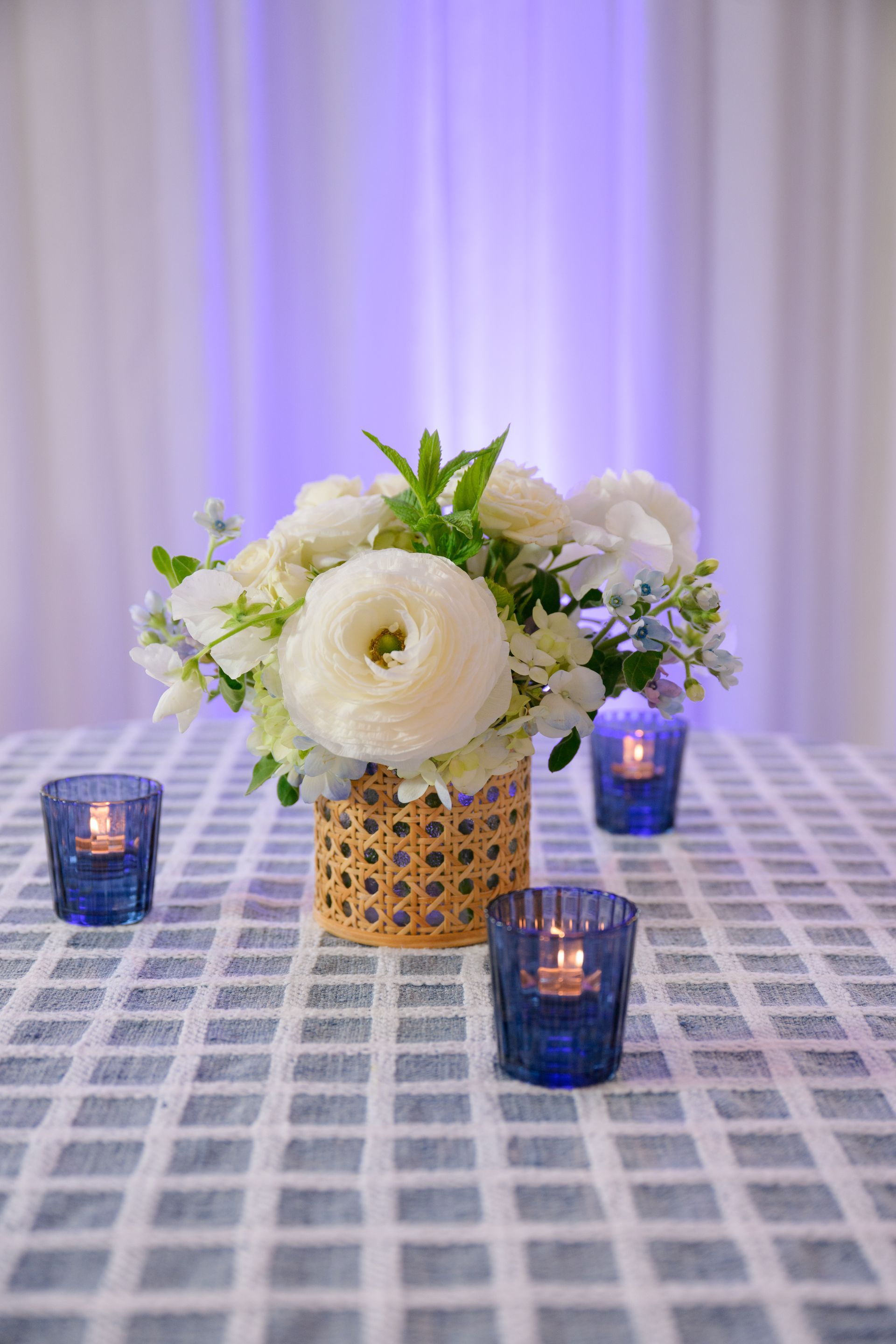 White floral centerpiece on a blue-checkered table, surrounded by lit blue votive candles; a blue-lit backdrop.