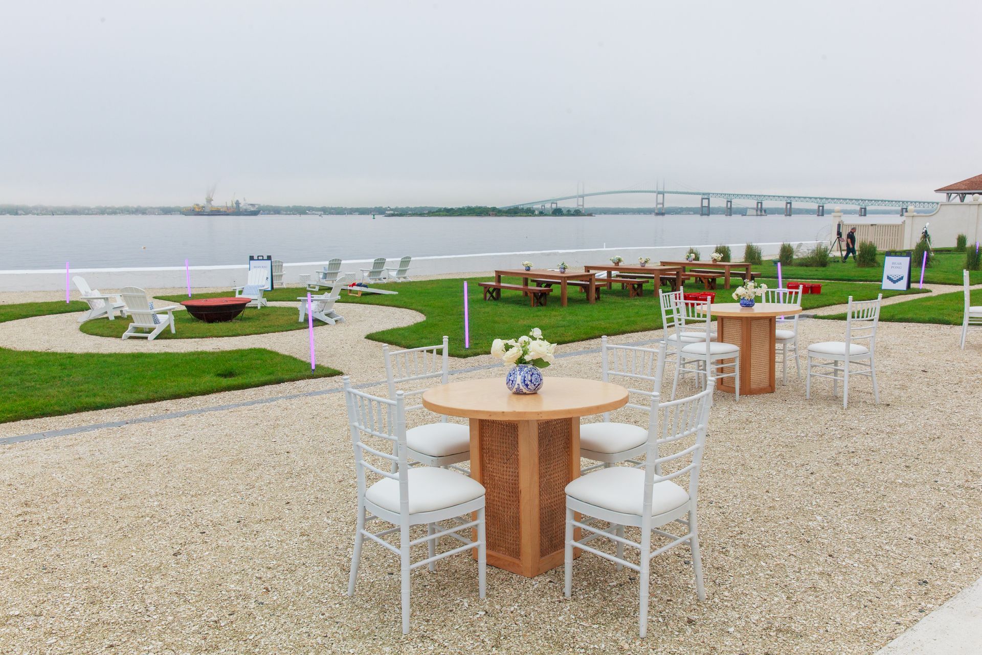 Outdoor seating area overlooking a beach and bridge, featuring round tables, white chairs, and green grass.