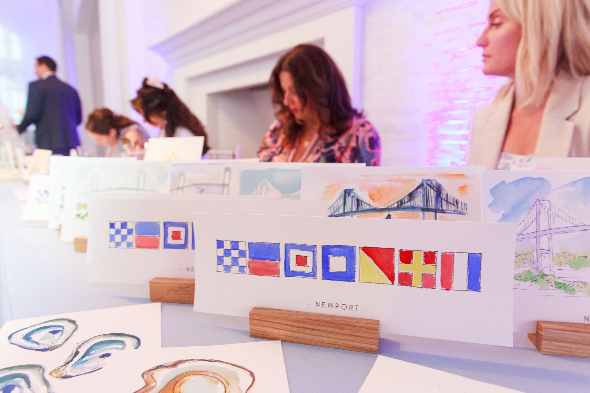 Watercolor art on a table with people in the background. Colorful nautical flag art and bridge sketches.