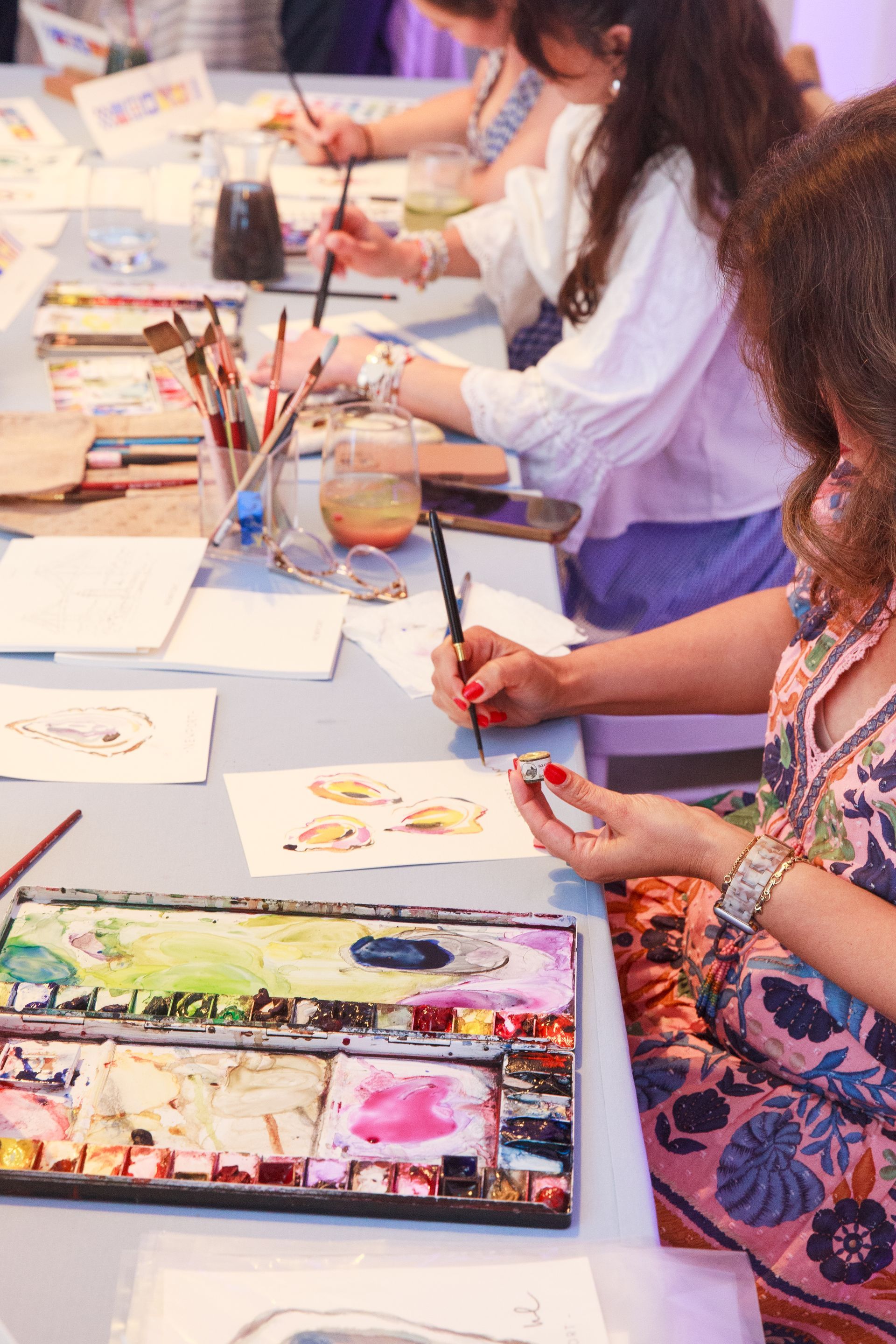 People painting with watercolors at a table, various colors and supplies visible, sunny day.