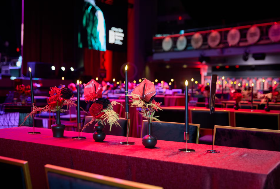 Formal dining tables set for event. Red tablecloths, centerpieces, lit black candles. Dark room.