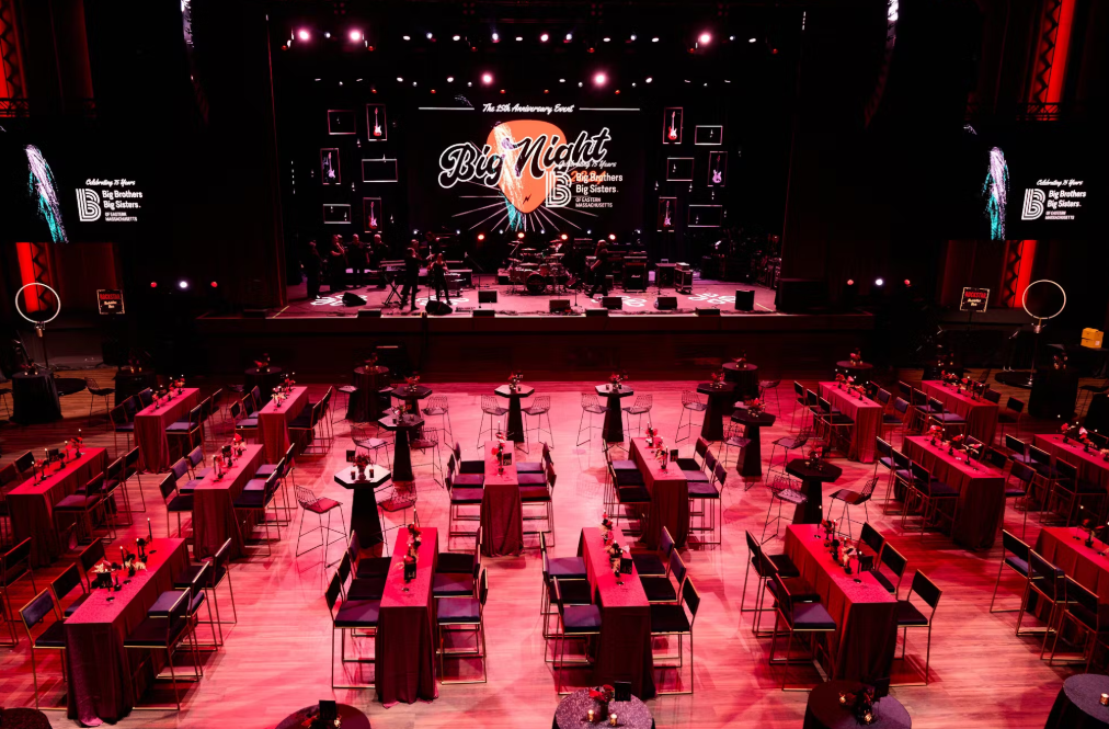 An event hall set for a gala. Tables with red linens face a stage with a band, bathed in red light.