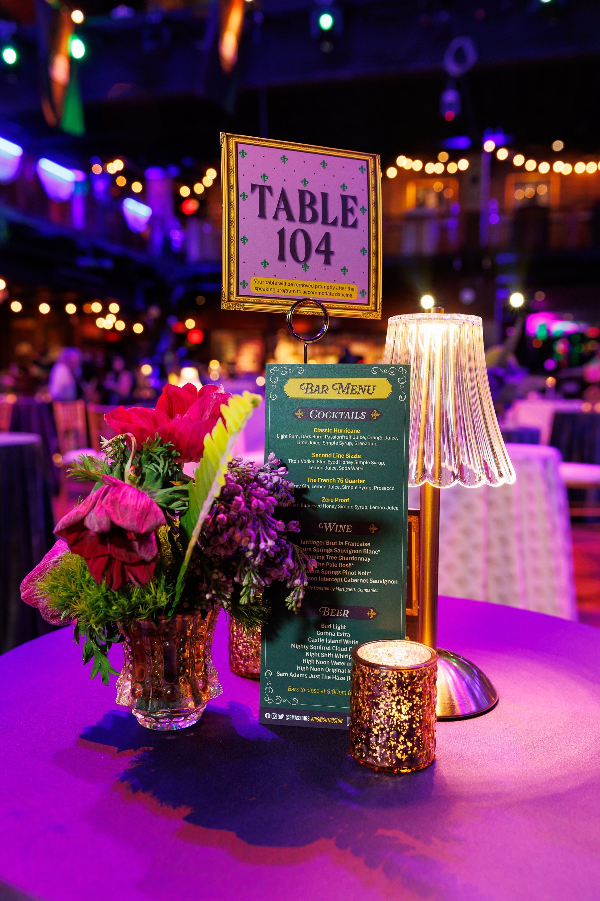 Table setting at event: floral arrangement, table number 104, menu, lamp, and candle on a purple tablecloth.
