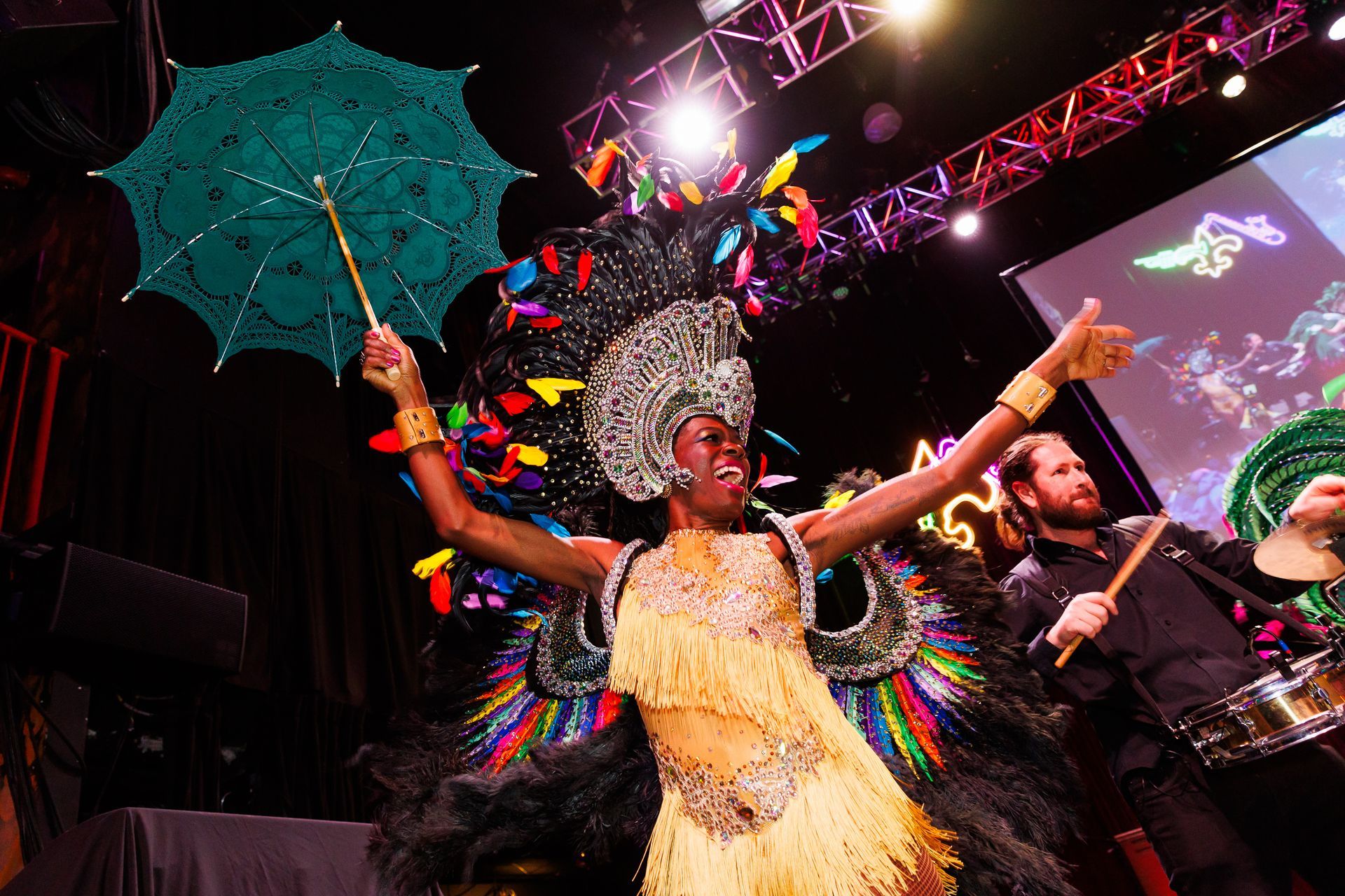 Woman in colorful Carnival costume with an open parasol, onstage; drummer behind her.