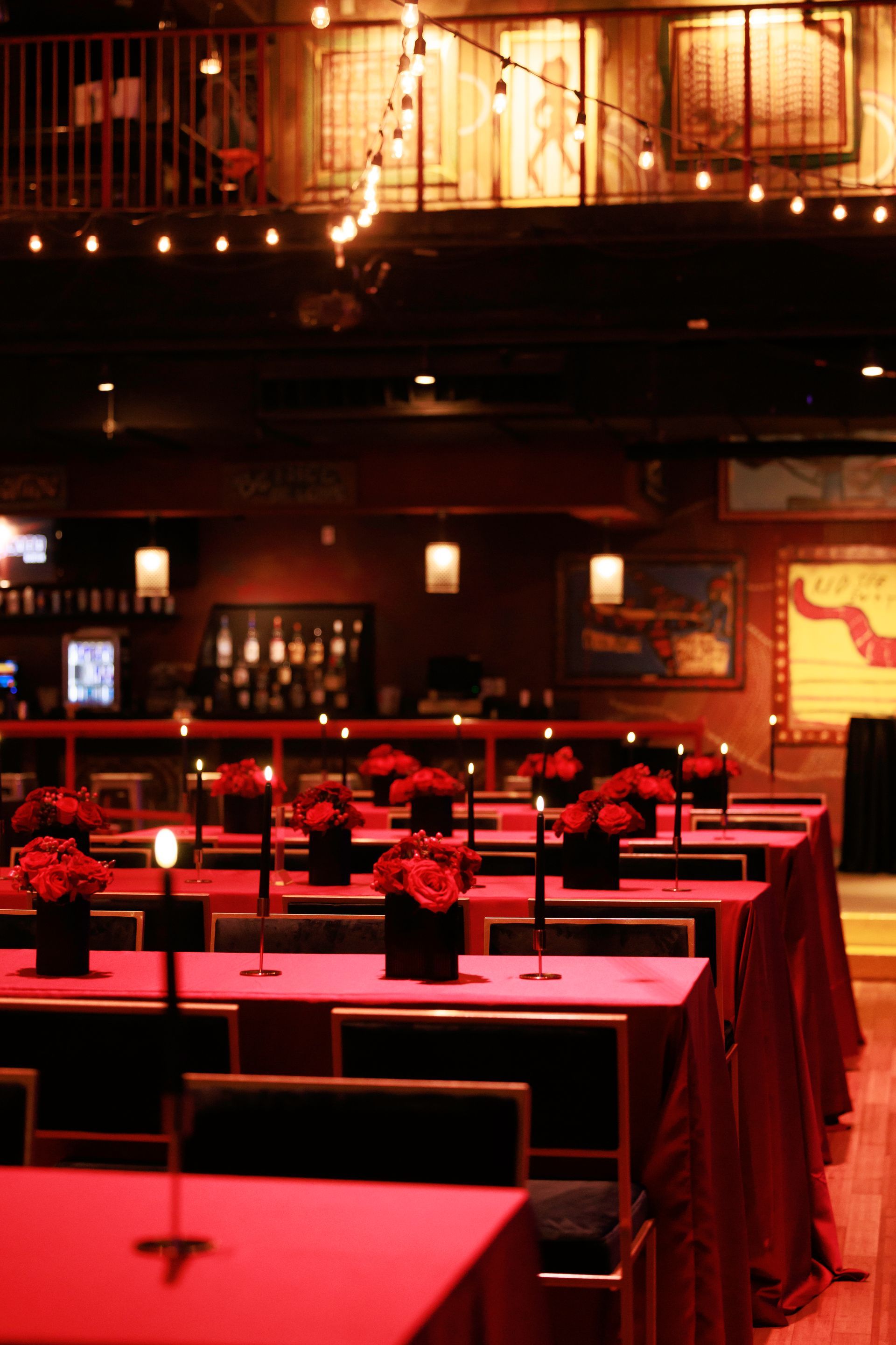 Rows of red-covered tables in a dark venue, set for a formal event. Flowers and candles adorn the tables.