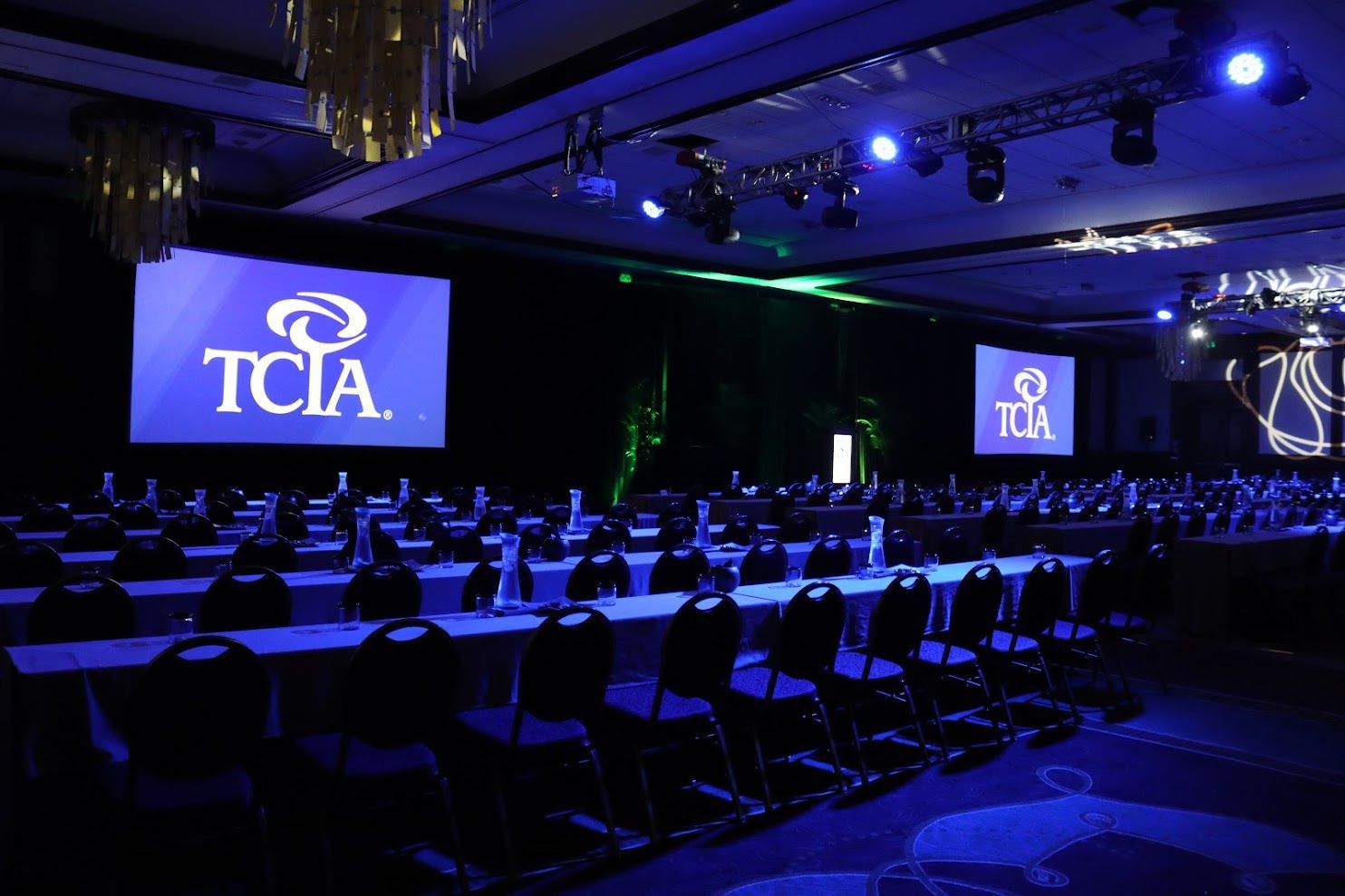 Conference hall with rows of chairs and tables, screens displaying the TCIA logo, and blue lighting.