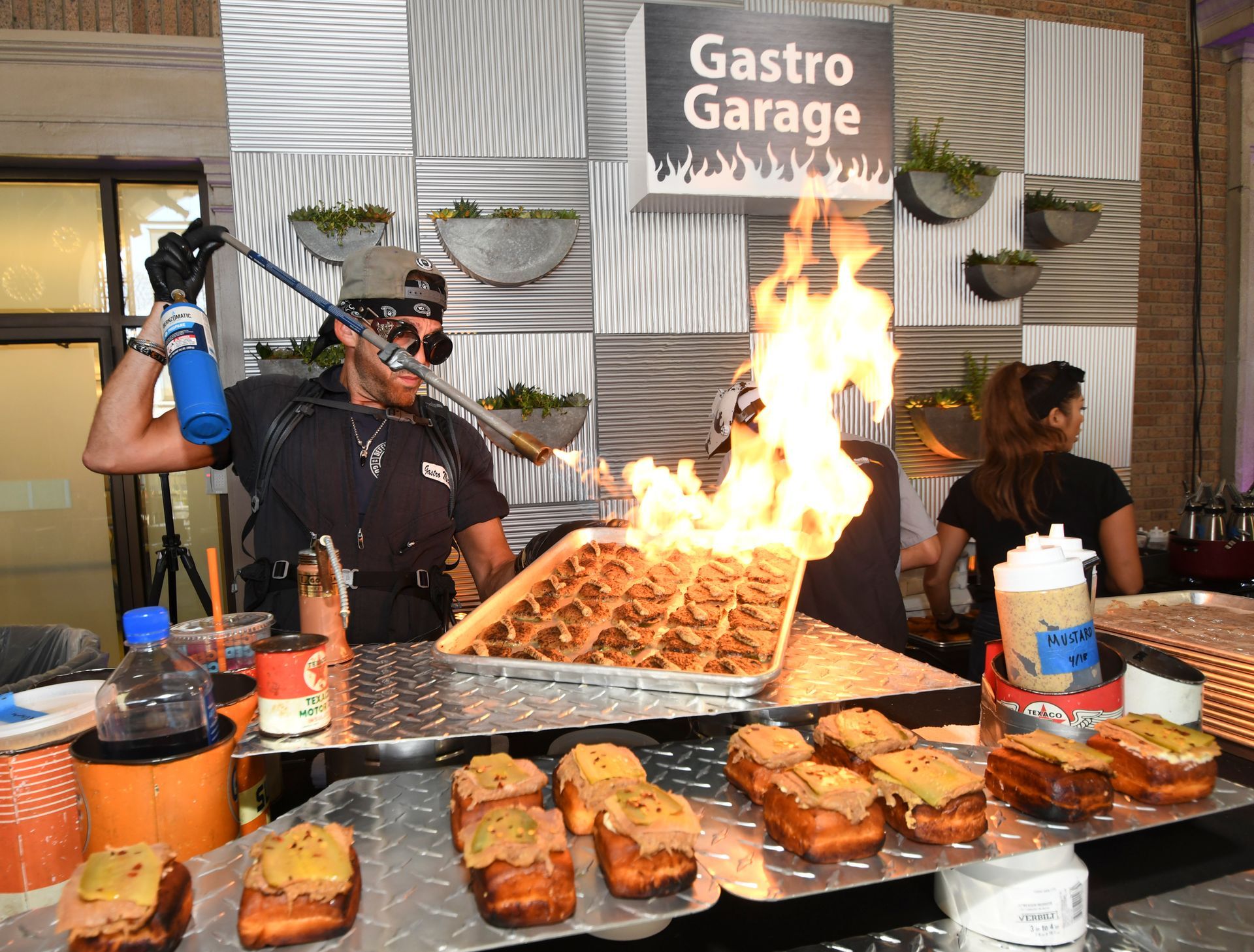 Chef using a torch to ignite food in a tray, at the Gastro Garage, next to finished desserts.